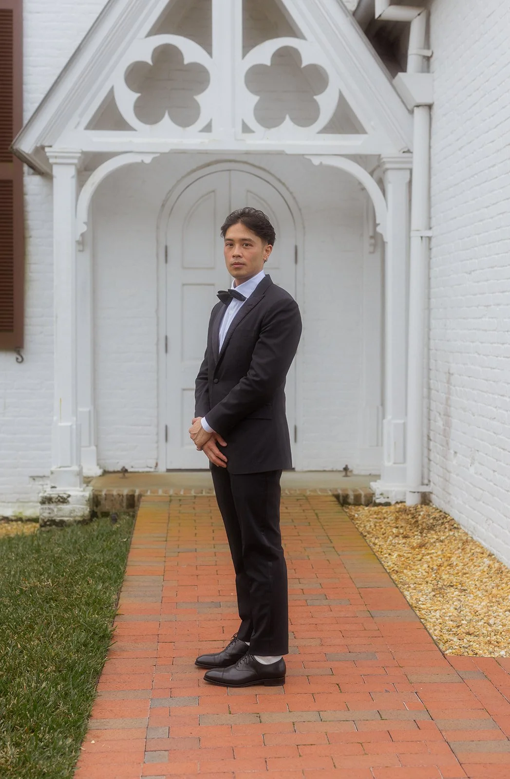 A man in a black tuxedo and bow tie standing on a brick pathway outside a white house with decorative trim and a closed door.