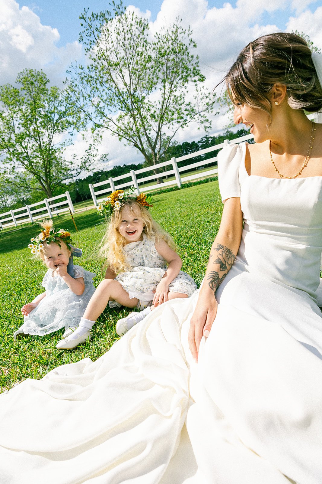 A woman in a white dress sitting on grass with two young girls, all wearing flower crowns, sharing a joyful moment outdoors under a partly cloudy sky.