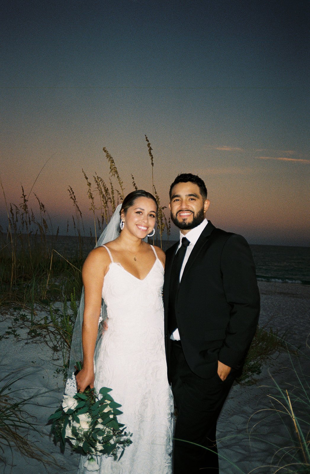 A newlywed couple standing on the beach at sunset, smiling for the camera. The bride is in a white wedding dress holding a bouquet, and the groom is in a black tuxedo with a white shirt. Behind them are tall grasses and the ocean, with a colorful sky