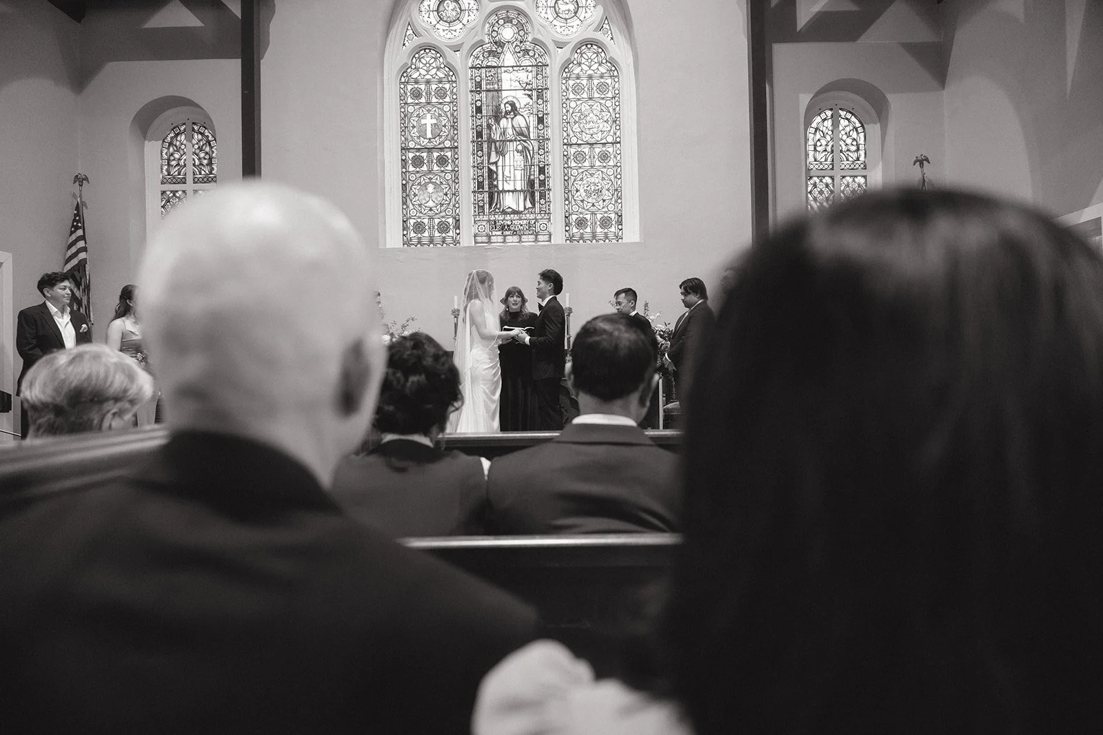 Black-and-white photo of a wedding ceremony inside a church, with the bride and groom exchanging vows at the altar, surrounded by guests seated in pews. The church has stained glass windows and an American flag is visible on the left side.