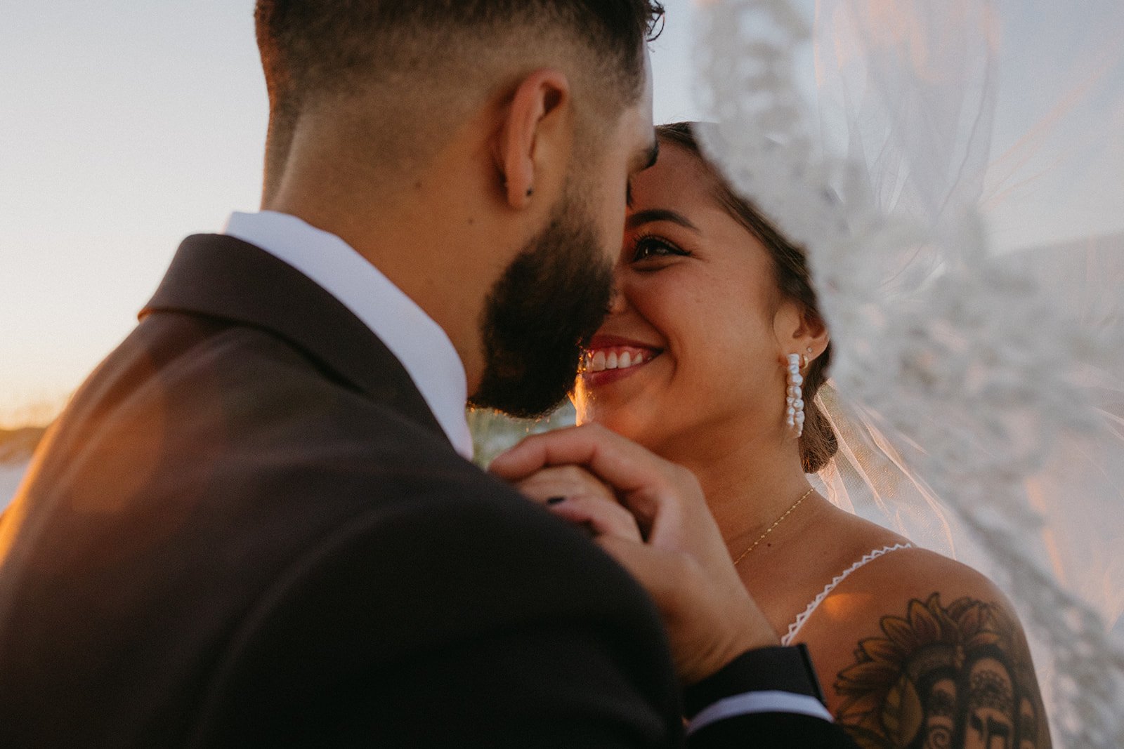 A close-up of a bride and groom smiling and holding hands, with their faces close together during a wedding ceremony outdoors at sunset.