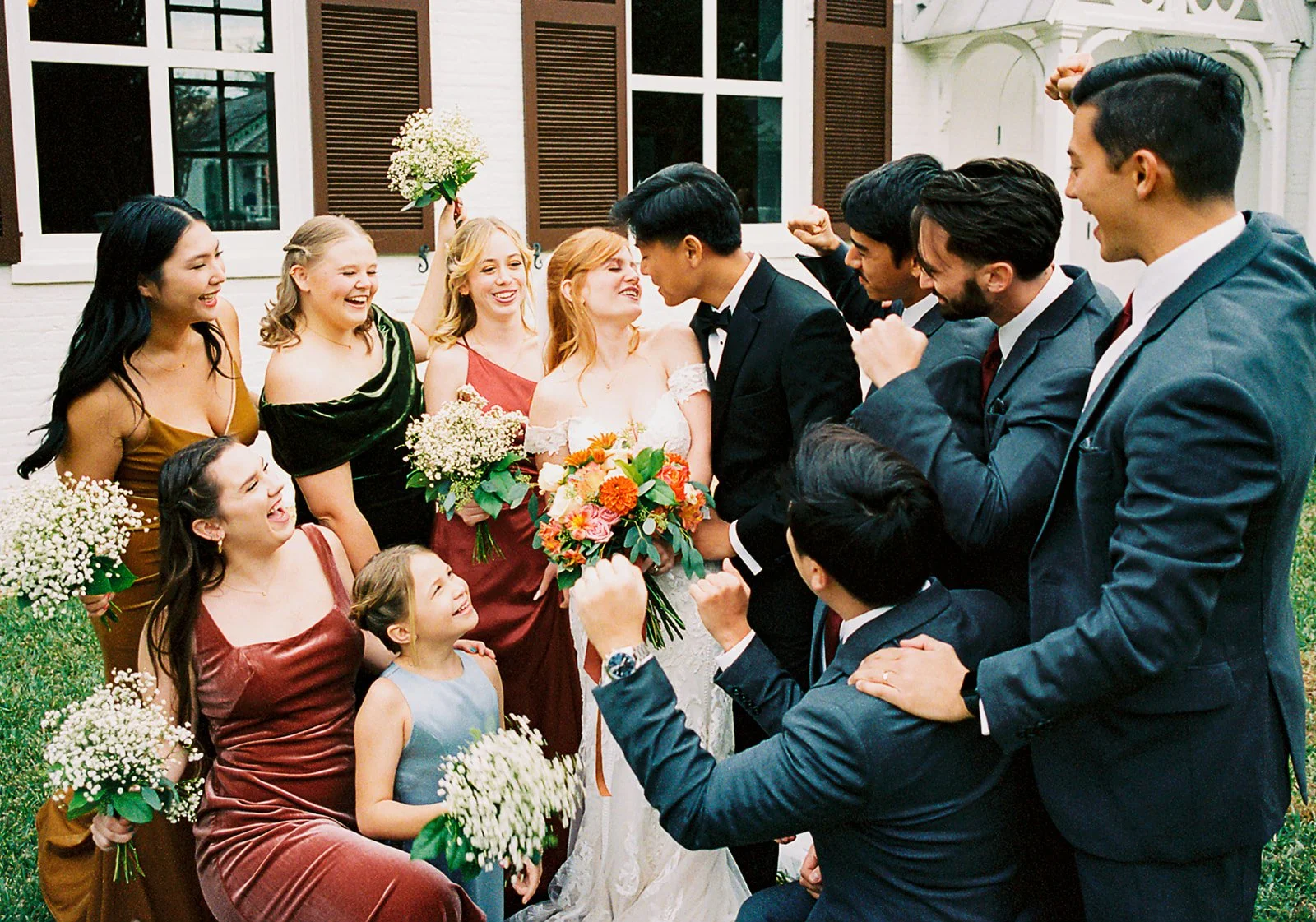 A diverse group of people in formal attire celebrating a wedding outside a house. The bride and groom are in the center, surrounded by friends and family holding flowers and cheering.