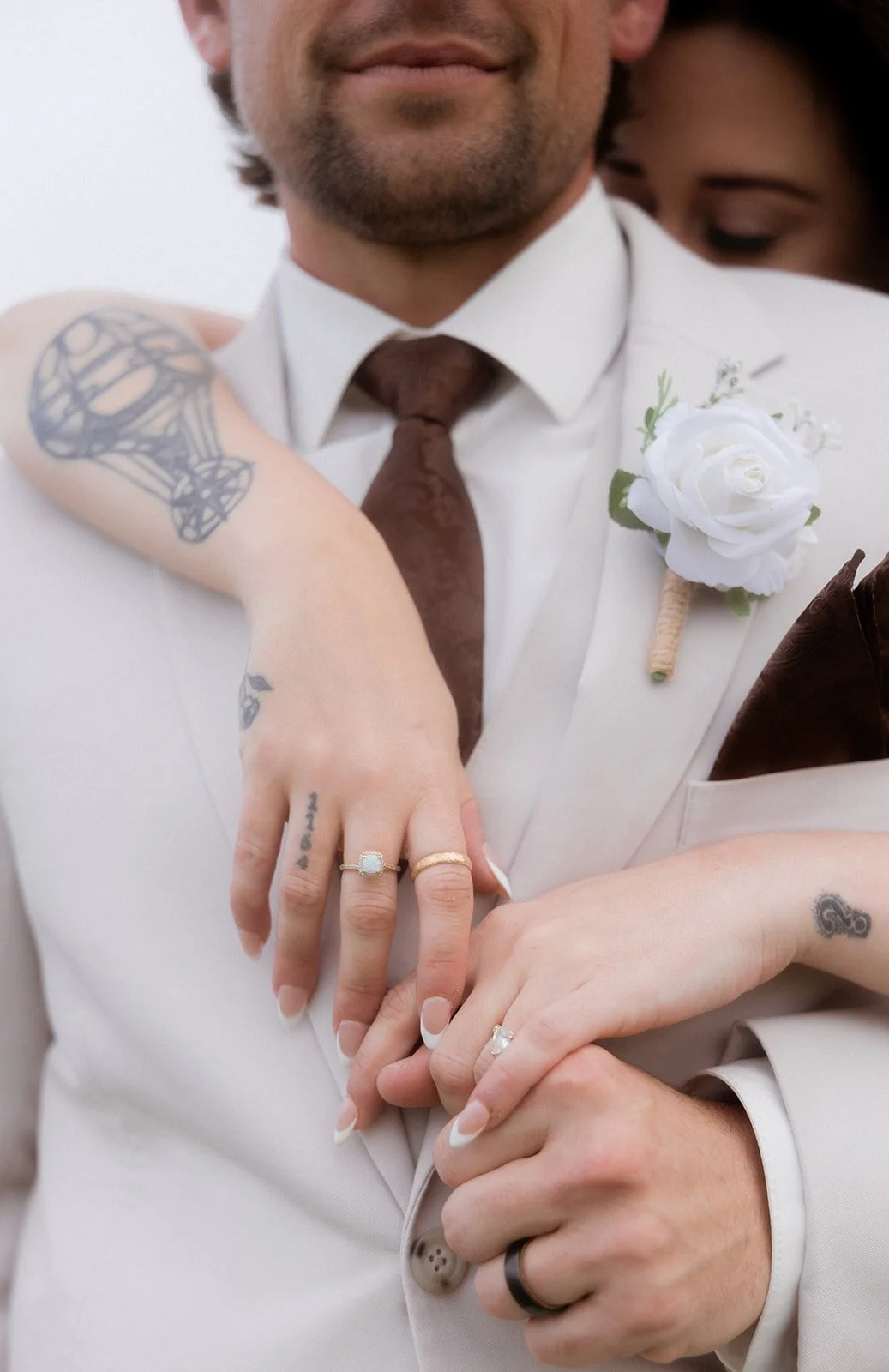 Close-up of a couple's hands showing wedding rings, with the woman's hand resting on the man's chest. The man wears a light-colored suit with a white shirt, brown tie, and a white boutonniere. The woman has tattoos on her arms and hands, and the back