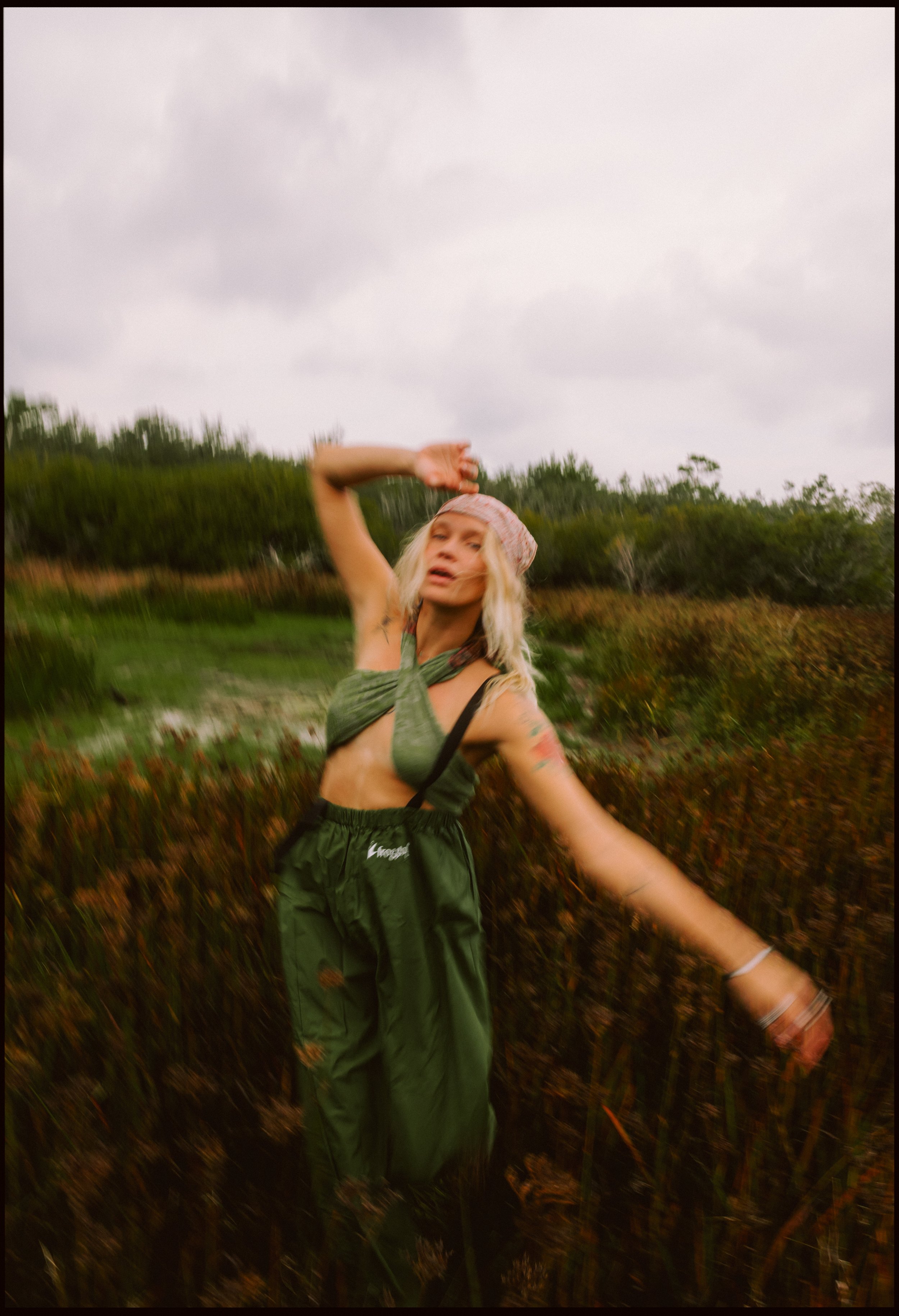 A woman with blonde hair wearing a pink bandana, green crop top, and green pants standing outdoors in a field of tall grass and plants during overcast weather.
