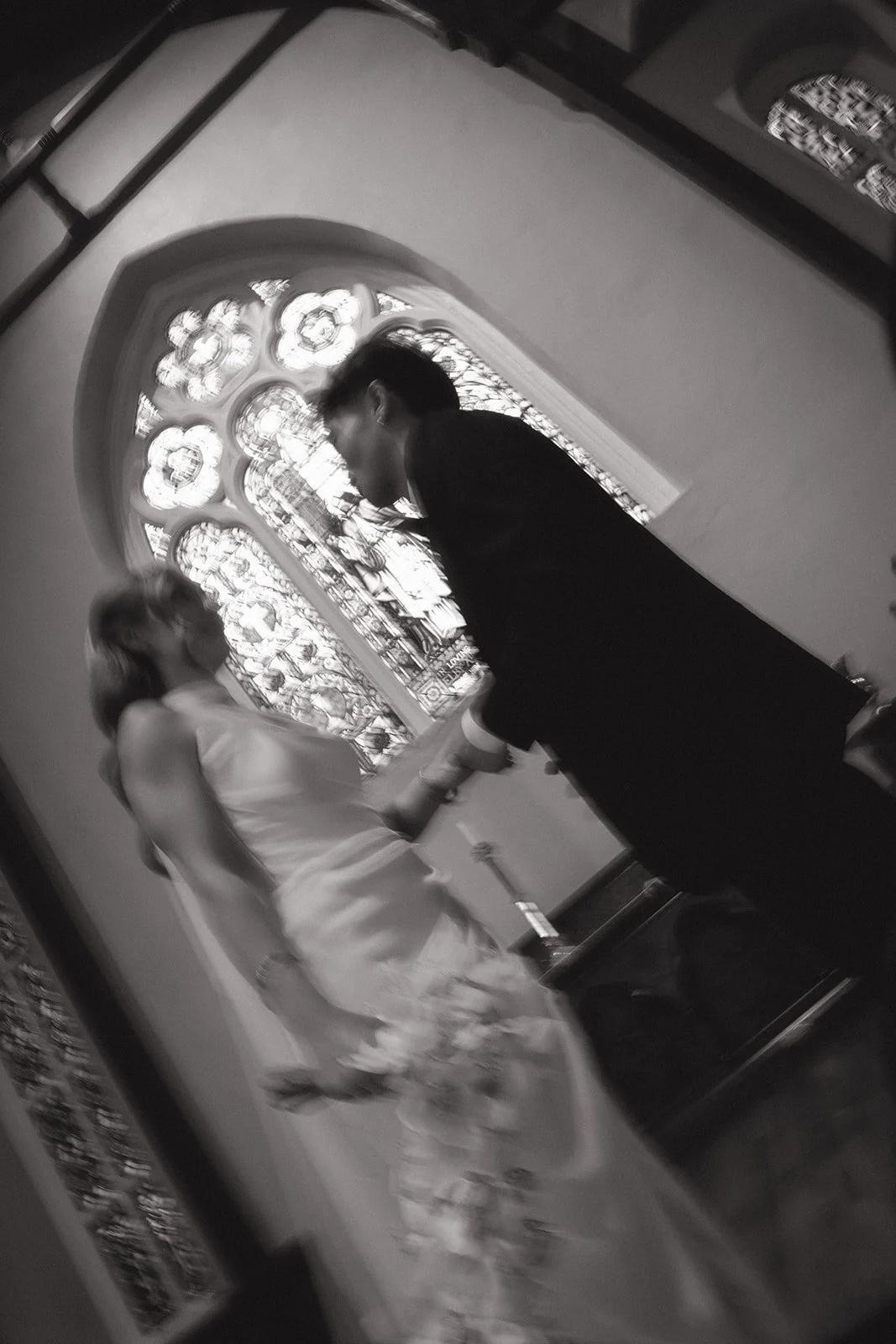 A bride and groom standing in front of a stained glass window inside a church, holding hands and facing each other. The photo is in black and white.