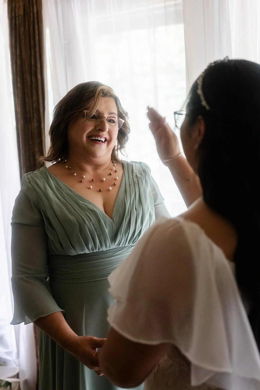 A woman in a green dress smiling at another woman with glasses, who is touching her face in an intimate moment indoors with natural light.