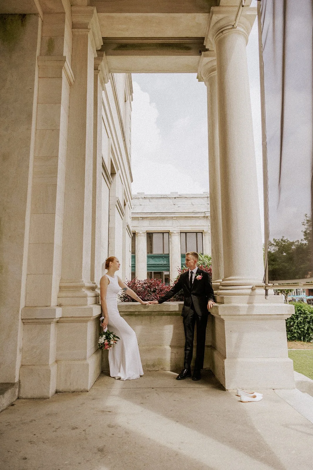 A bride in a white wedding dress and a groom in a black suit and tie holding hands and looking at each other under large stone columns with an old building and greenery in the background.