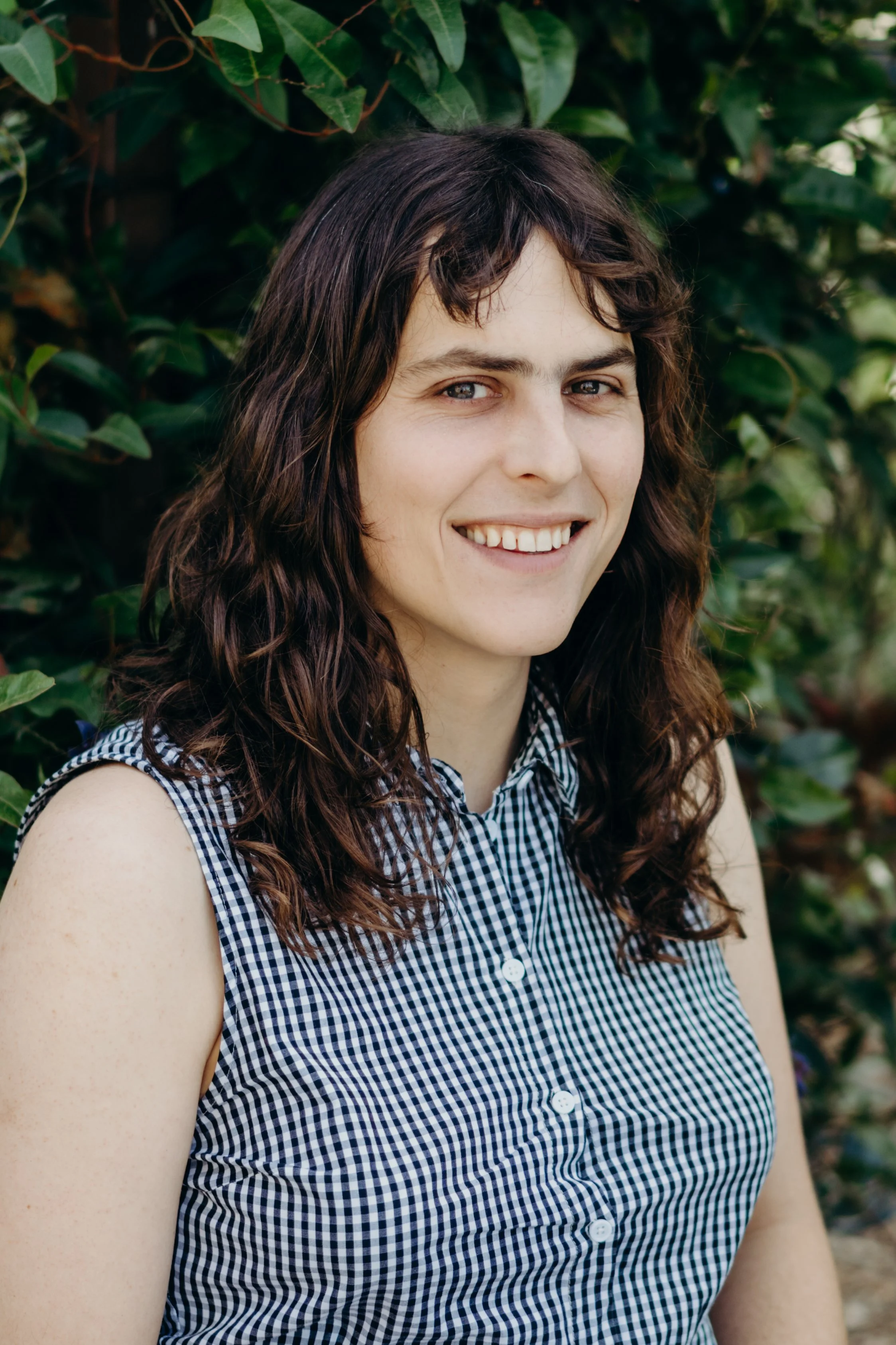 A woman with wavy brown hair smiling in front of green bushes, wearing a sleeveless, button-down checkered shirt.