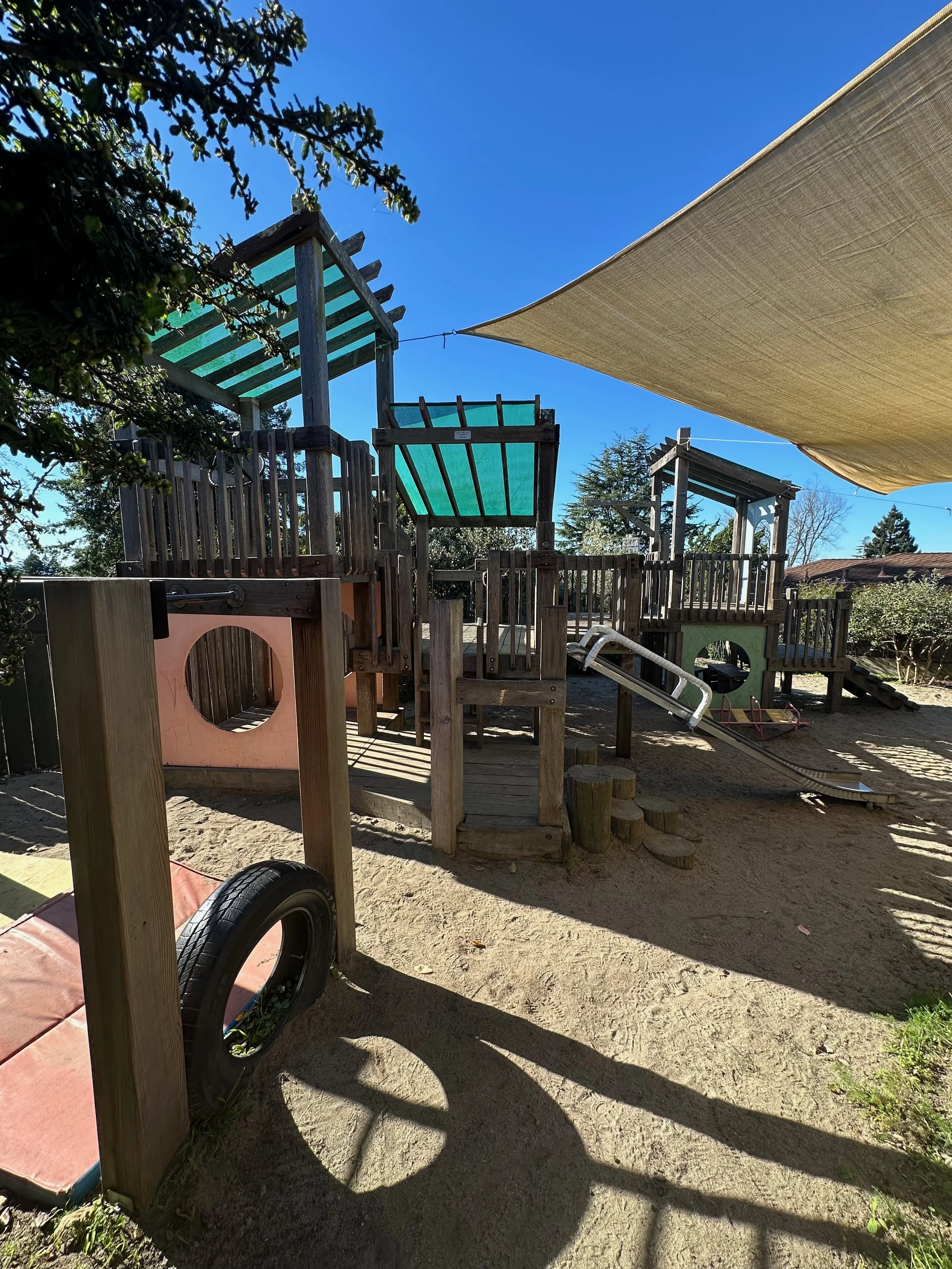 Wooden playground structure with slides, climbing areas, and shaded canopy on sandy ground outside.