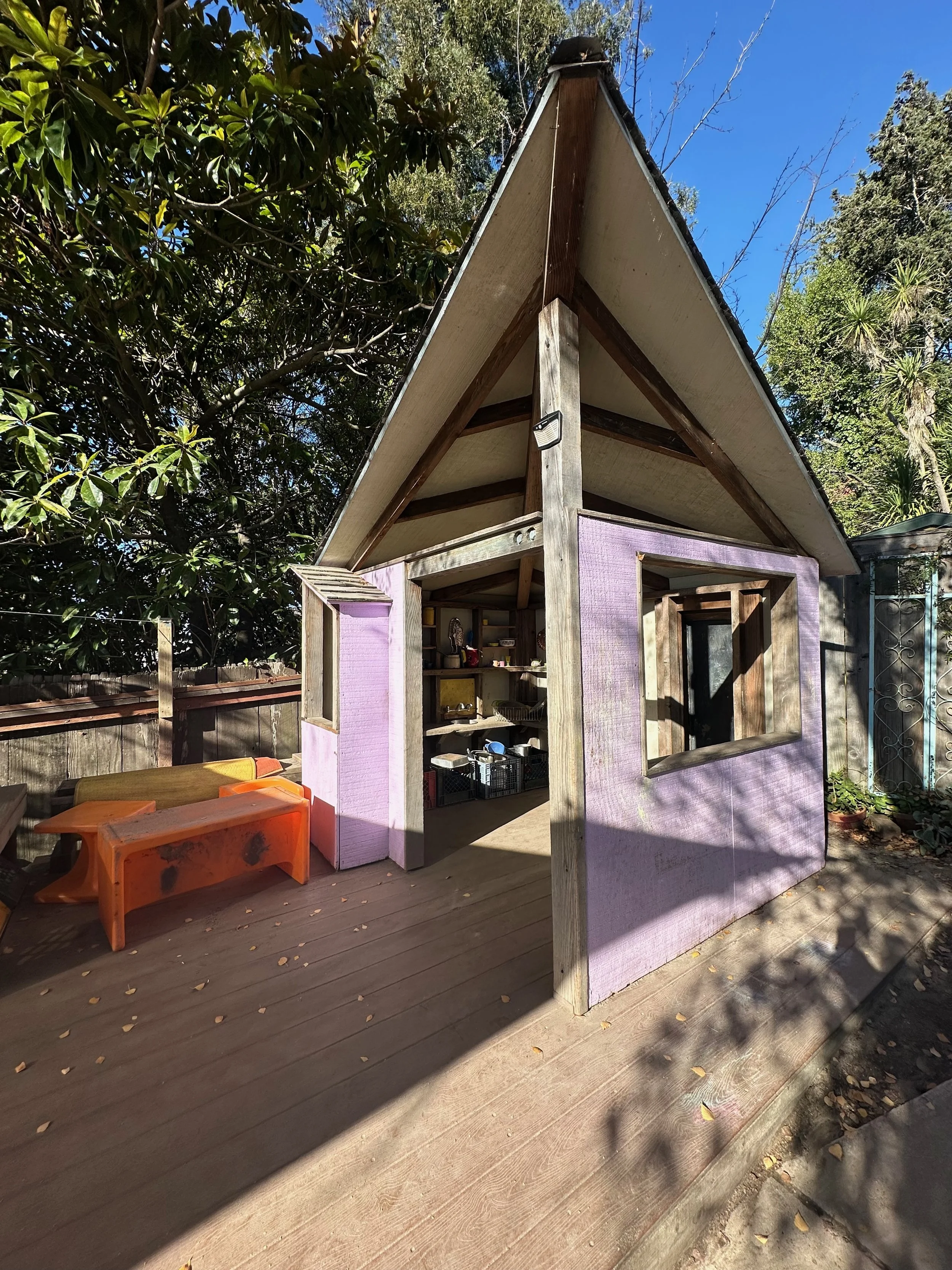 A small purple playhouse with a peaked roof, open front, and window, situated on a wooden deck surrounded by trees.