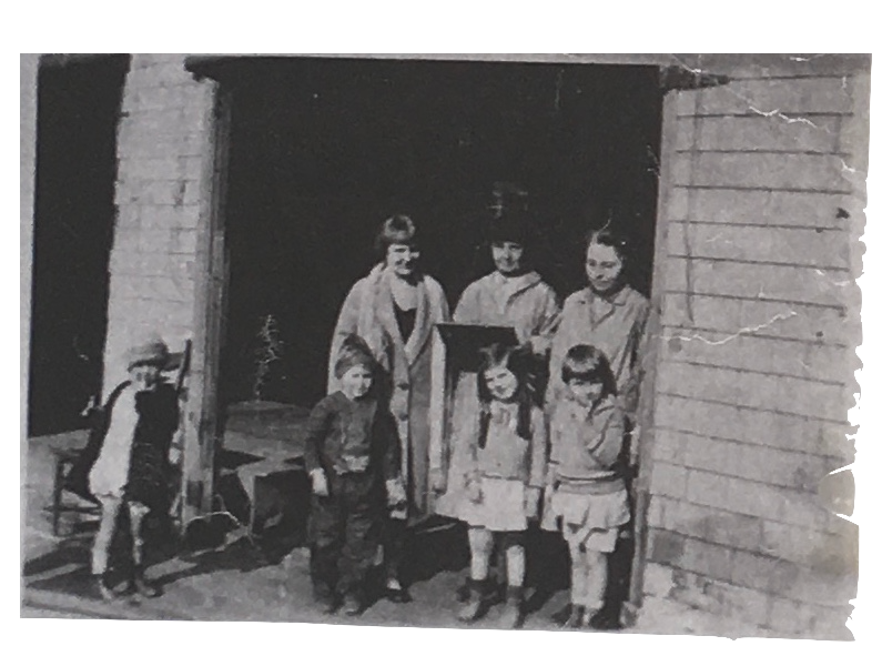 A vintage black-and-white photo of six children and one adult standing outside a wooden building. The children are dressed in old-fashioned clothing, with some wearing dresses and others in pants and shirts. The adult is holding a book or a paper. There is a wooden chair nearby and some outdoor plants.