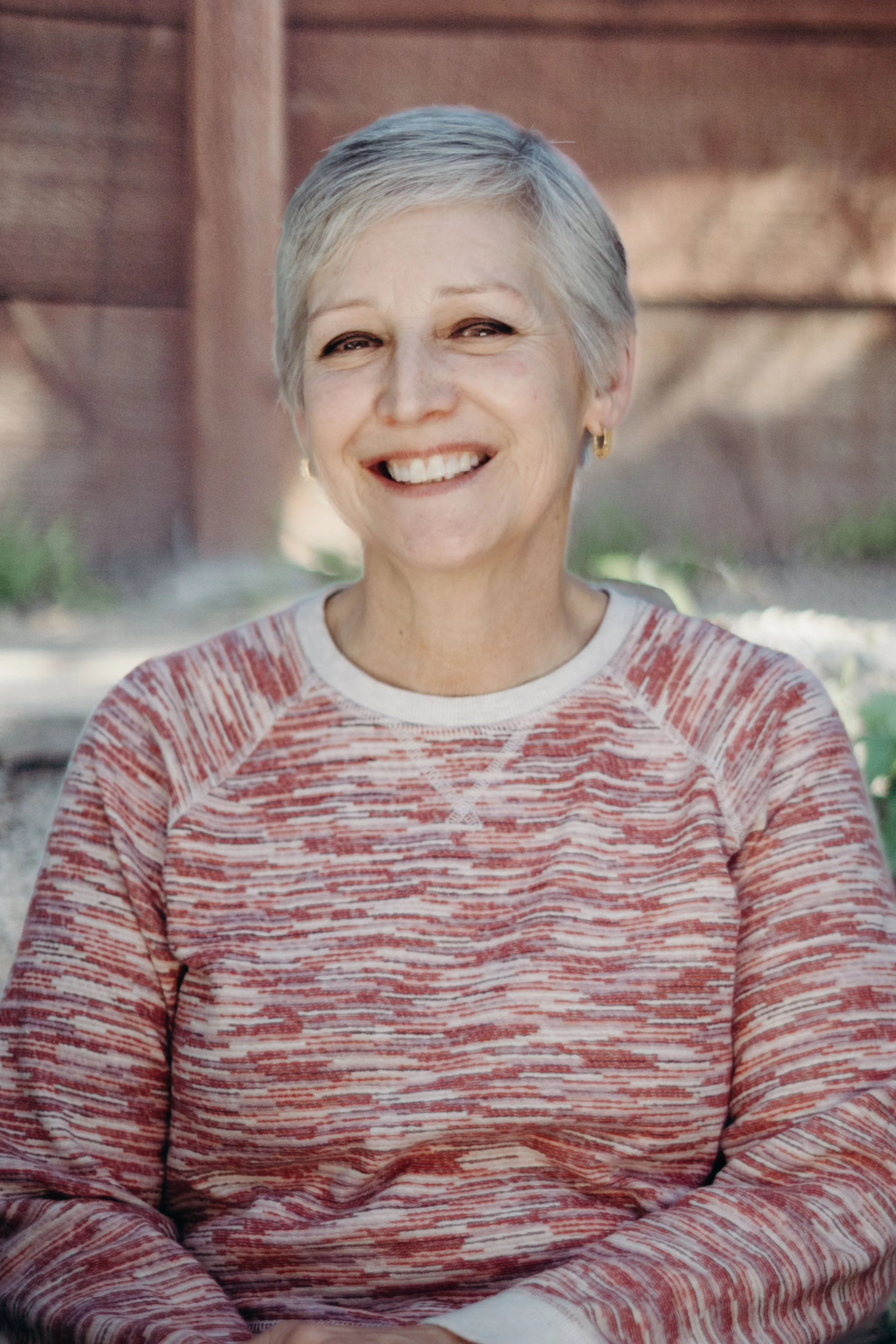 A smiling older woman with short gray hair, wearing a red and white patterned sweater, sitting outdoors with a wooden background.
