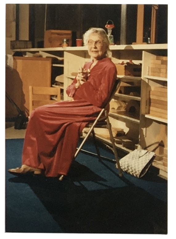 An elderly woman with gray hair wearing a red dress, sitting on a folding chair with her hands clasped, in a room with wooden shelves and boxes.