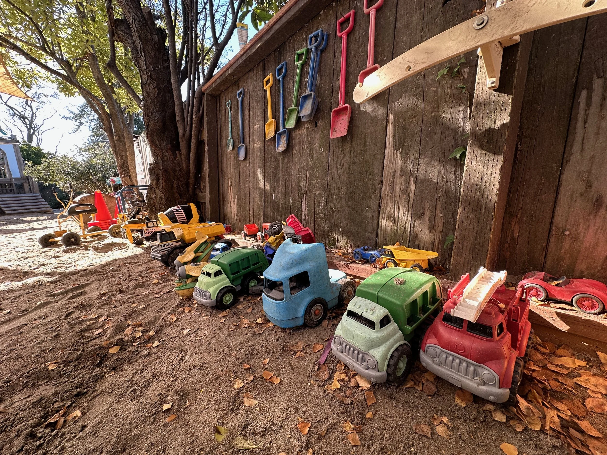 Colorful children's toy trucks lined up on dirt ground next to a wooden fence with plastic shovels hanging on it, and a tree with leaves providing some shade.