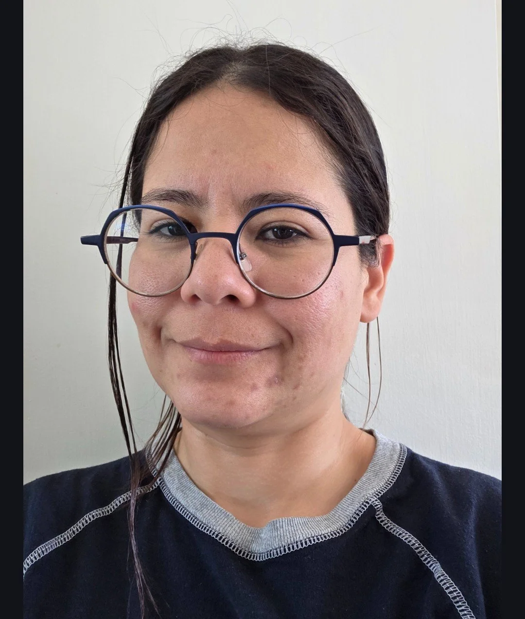 A woman with dark hair, wearing round glasses and a navy blue shirt, smiling in front of a plain white wall.