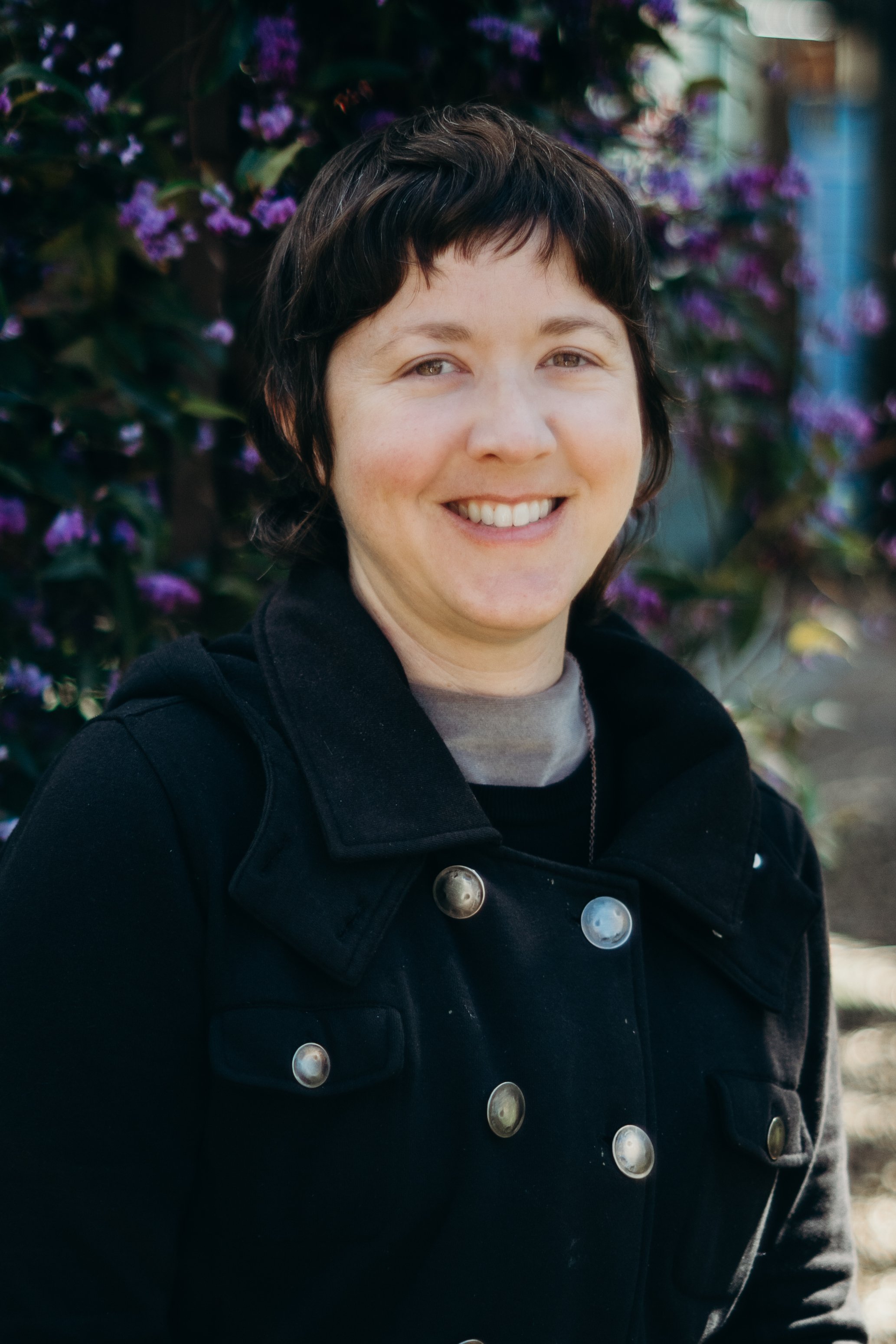 A woman with short dark hair smiling outdoors, standing in front of purple flowering bush, wearing a black jacket.