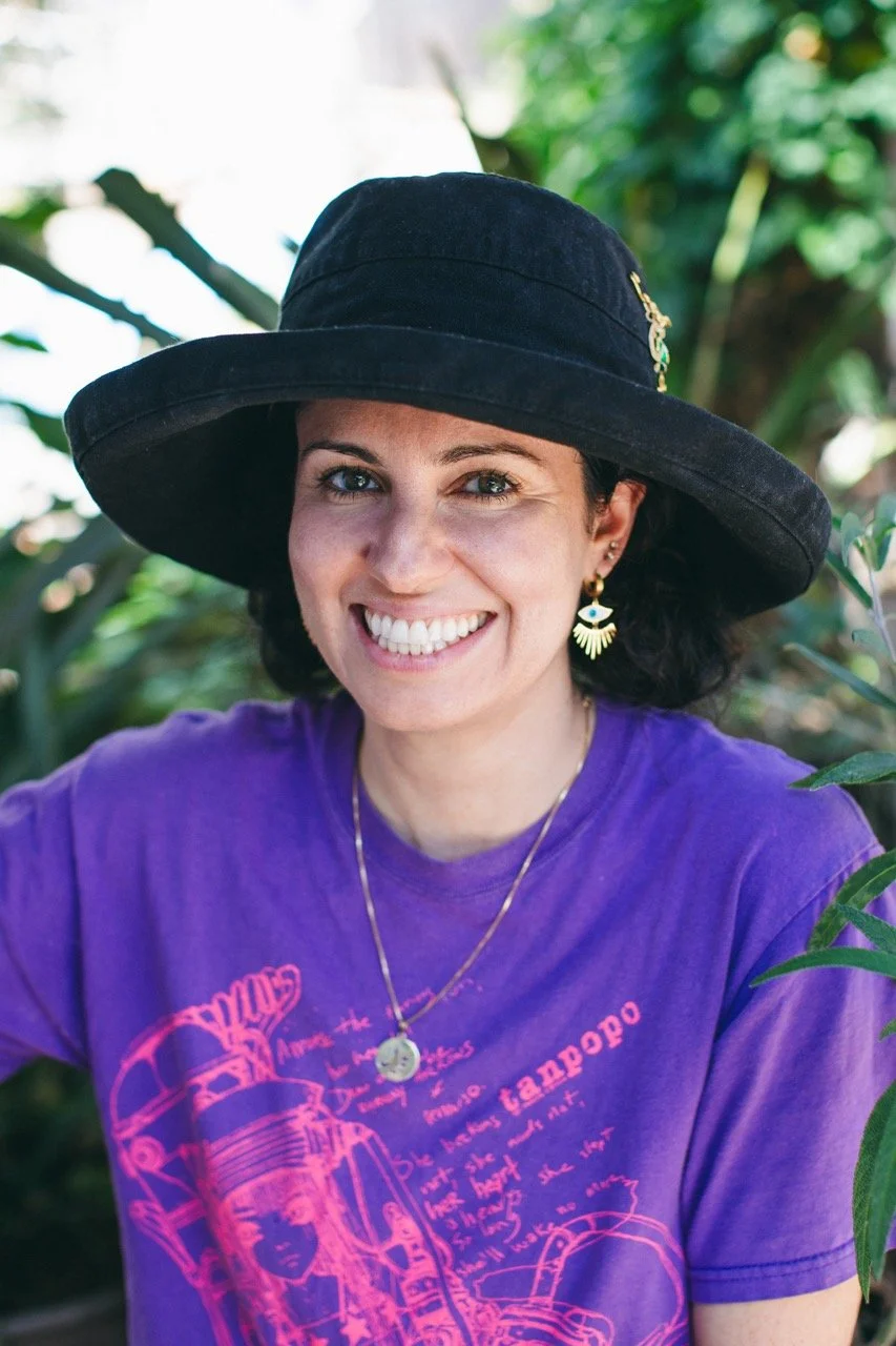 A woman smiling outdoors wearing a black wide-brimmed hat, purple graphic t-shirt, gold jewelry, and earrings with eye designs, surrounded by green plants.