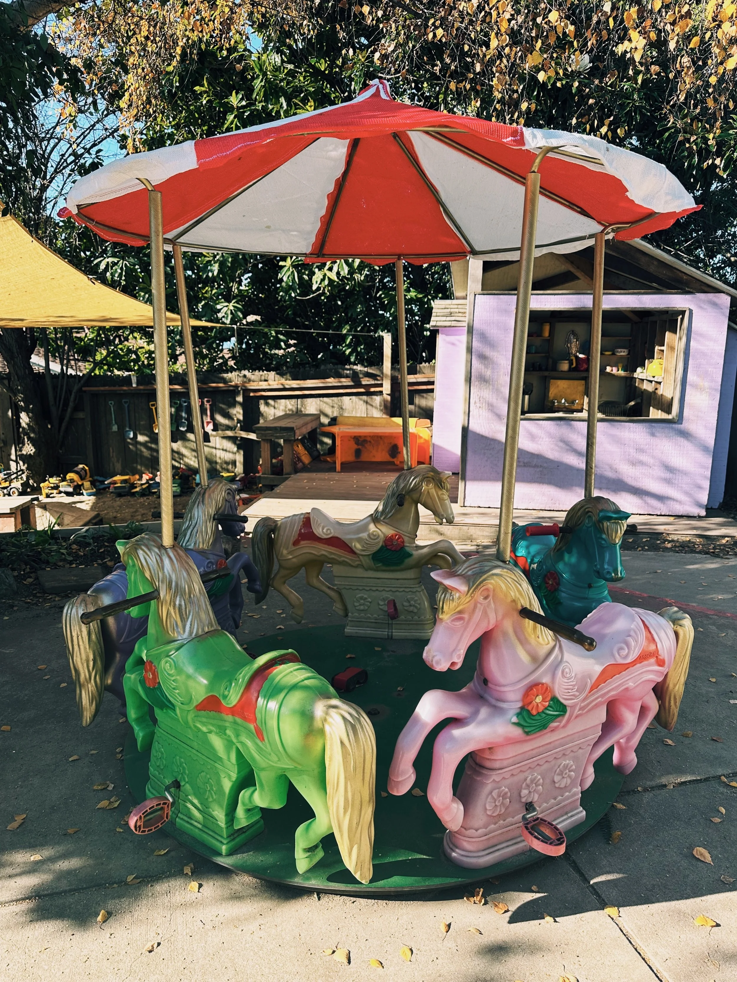 Colorful kiddie carousel with pastel pink, green, purple, and blue horses under a large red and white umbrella in a backyard play area.