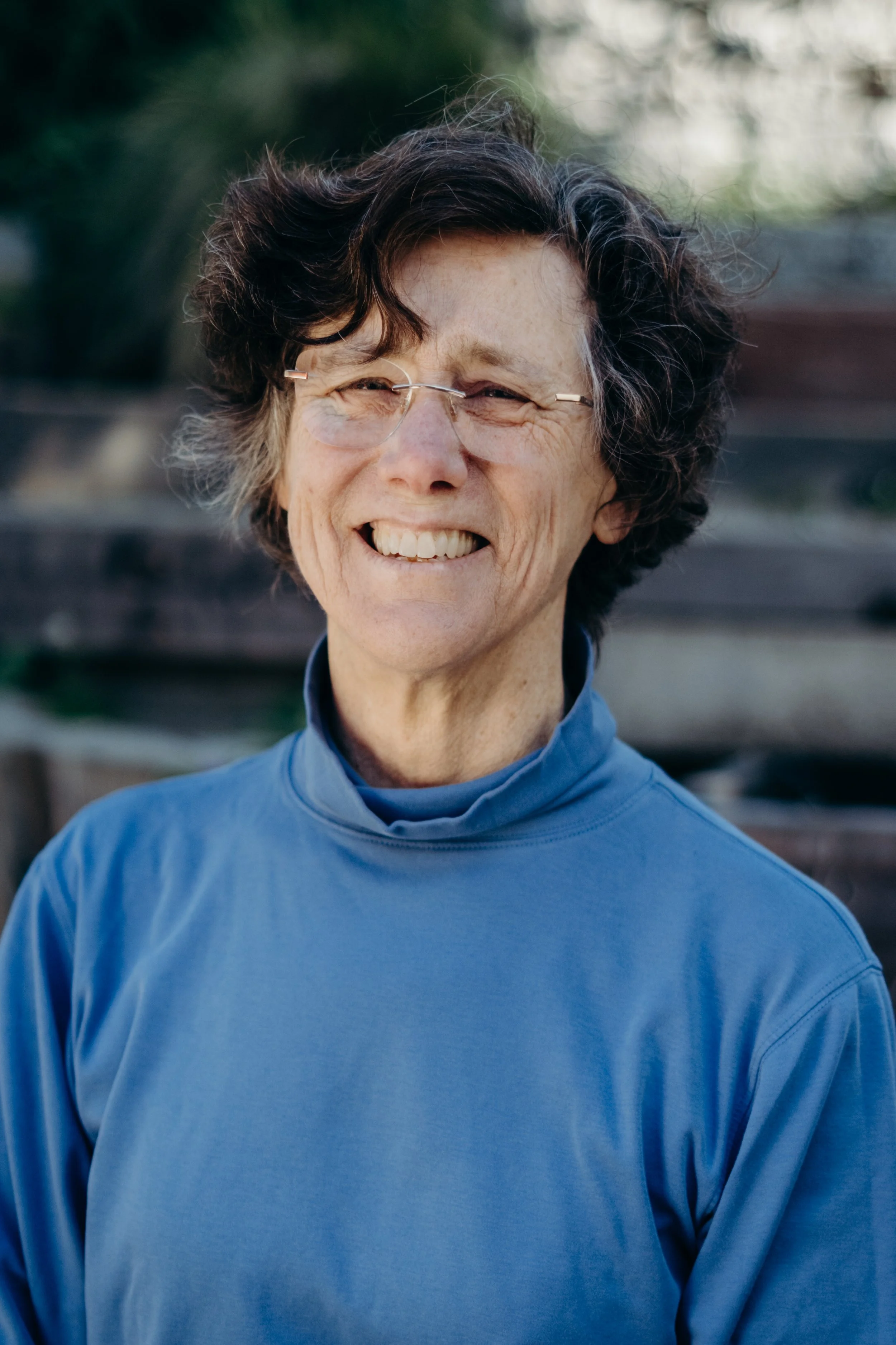 Smiling person with glasses, wearing a pink puffy vest over a gray shirt, sitting outdoors in front of a painted wooden fence.