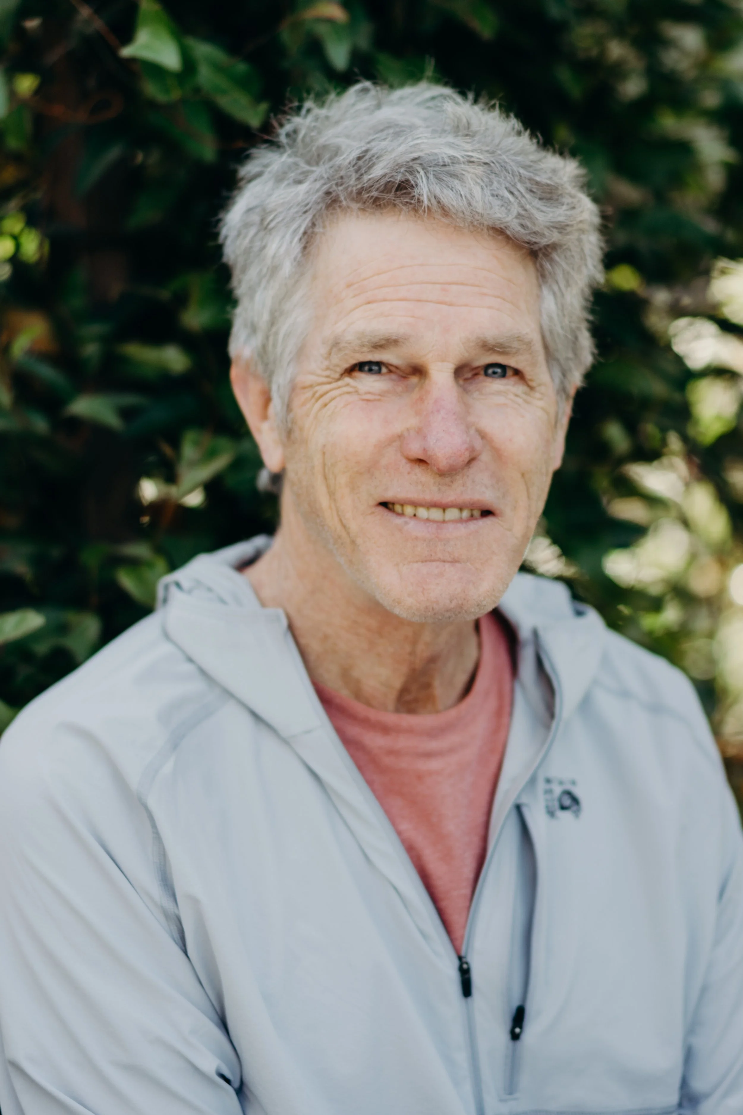 A portrait of an older man with gray hair and blue eyes, smiling slightly, outdoors in front of green foliage, wearing a light gray jacket over a reddish-pink shirt.