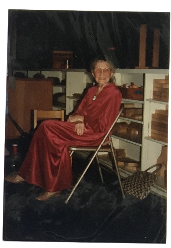 An elderly woman with glasses and gray hair, wearing a red dress, sitting on a folding chair inside a room with shelves filled with bricks and other items, smiling at the camera.