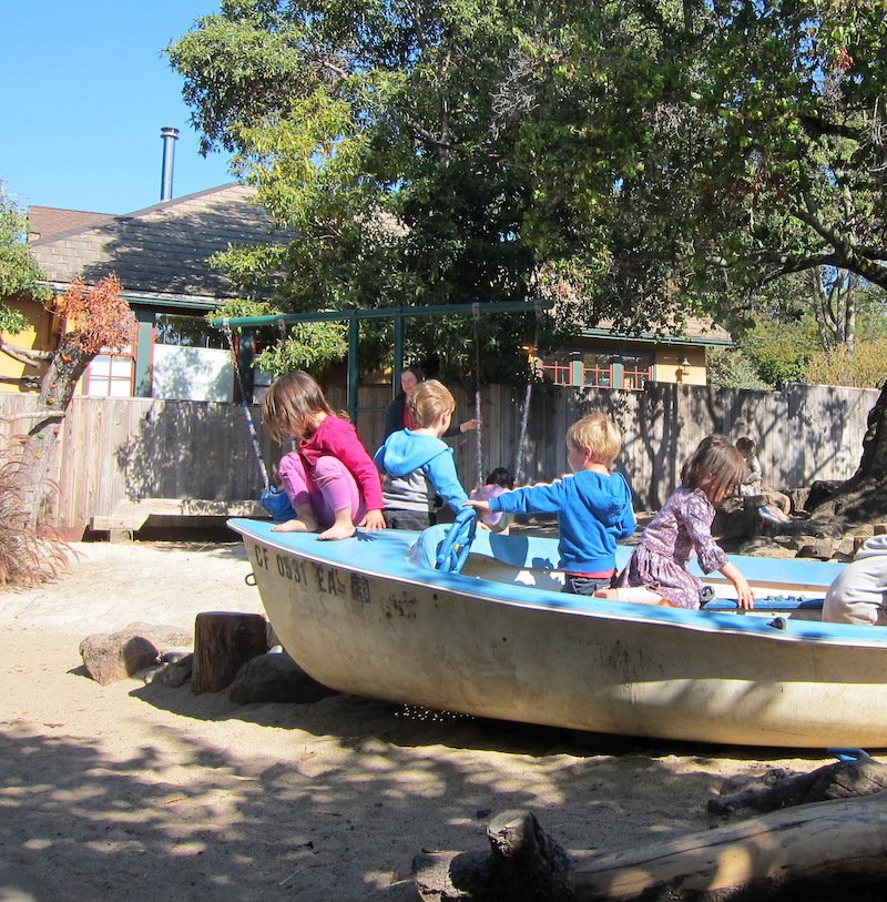 Children playing in a boat on a playground with trees and houses in the background.