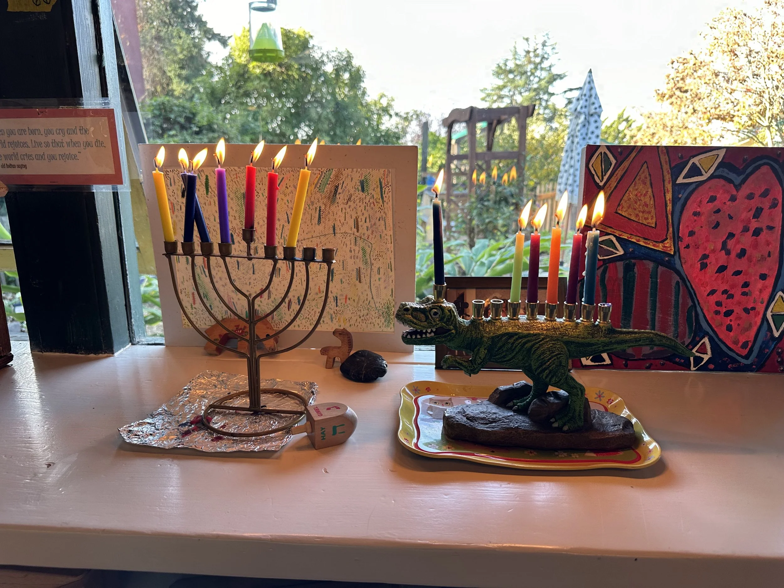 A Hanukkah menorah with lit candles and a Toy Story Rex figure with lit candles on its back, surrounded by children's drawings and decorations on a windowsill.