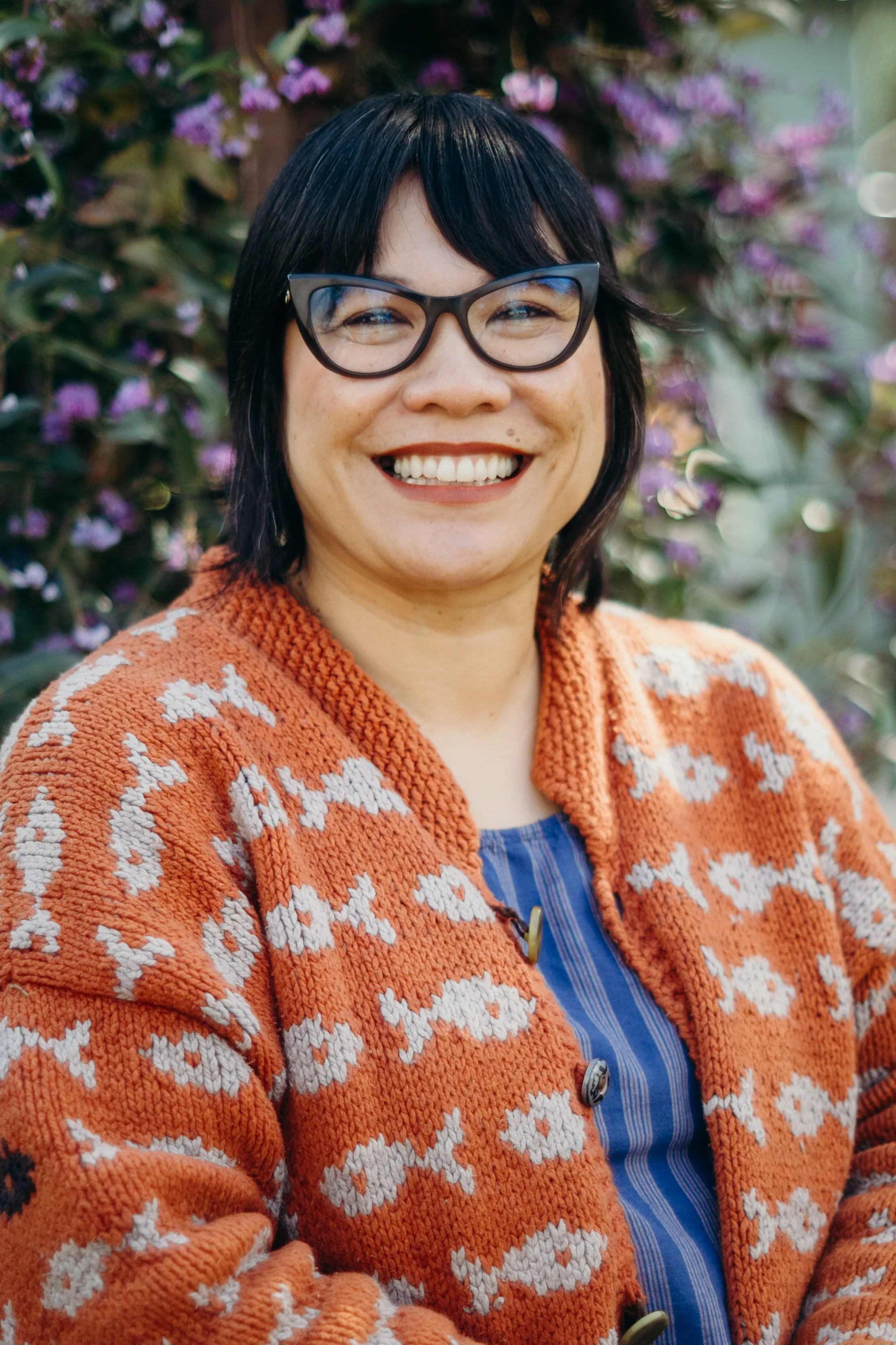 A woman with dark hair, glasses, and a warm smile wearing an orange and white patterned cardigan standing outdoors with purple flowers in the background.