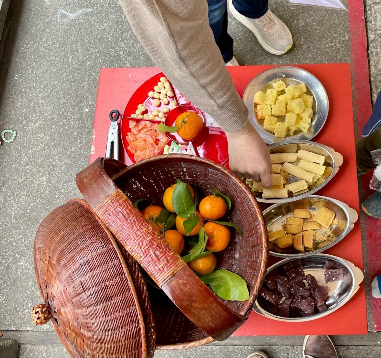 A person reaching for some food on a red table with various traditional Asian sweets, fruits, and snacks displayed in metal and plastic containers.