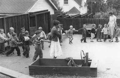 Children holding hands and walking in a line outside, accompanied by a woman, with a toy car in the foreground.