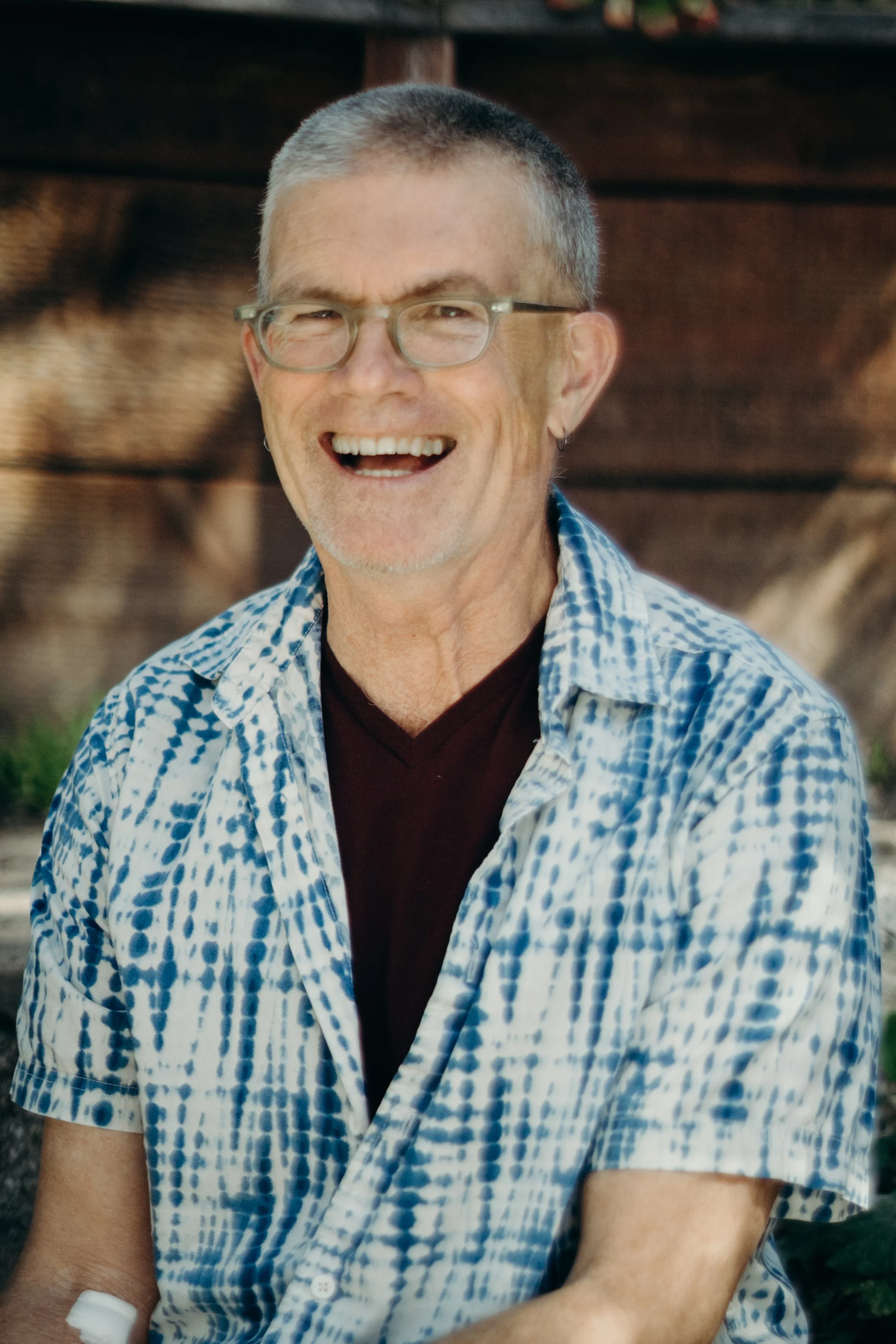 A smiling man with short gray hair, glasses, and a beard, wearing a blue and white tie-dye shirt over a dark t-shirt, standing outdoors in front of a wooden background.