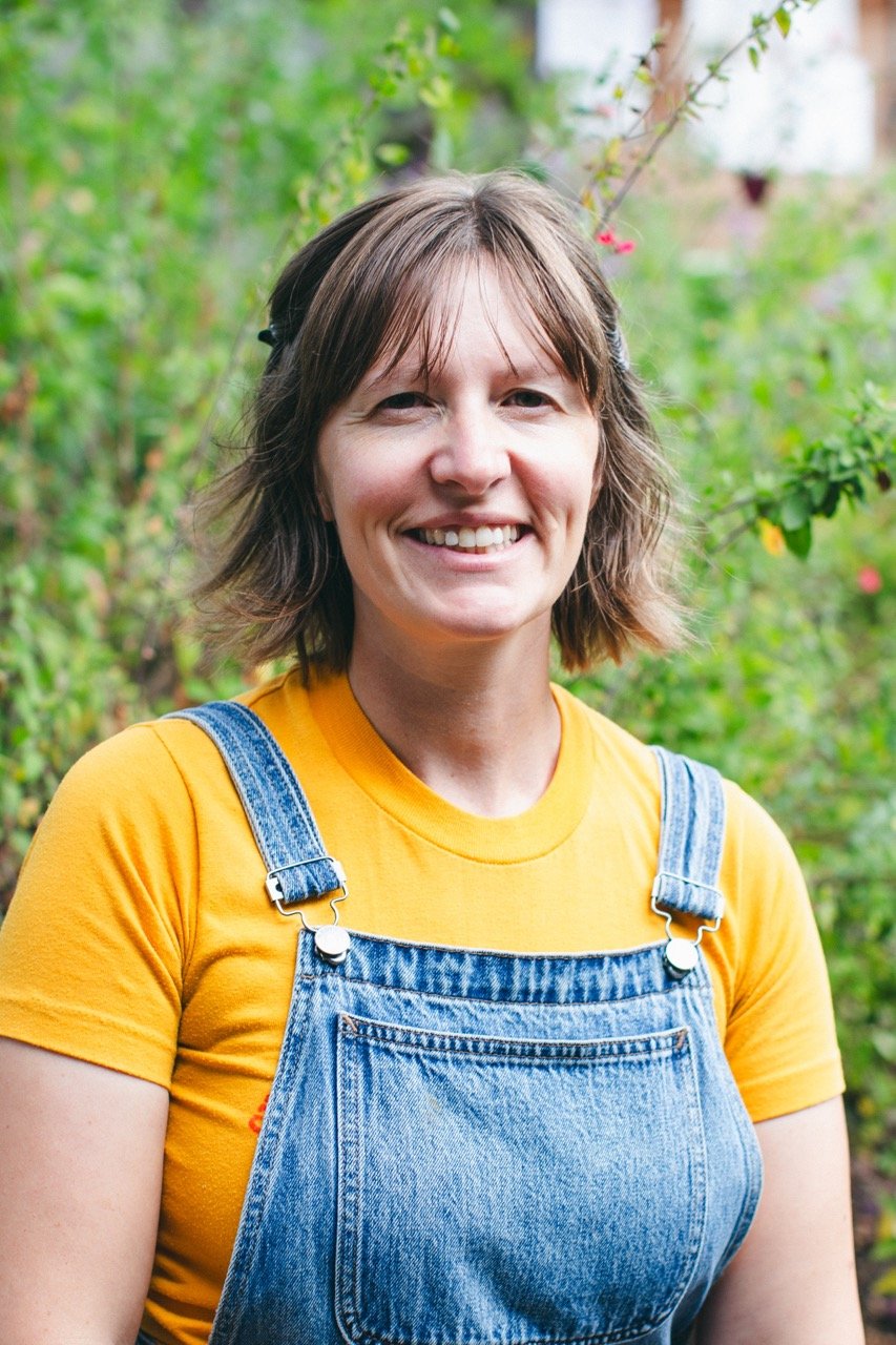 A woman with shoulder-length brown hair smiling at the camera, wearing a yellow T-shirt and denim overalls, standing outdoors with green foliage in the background.