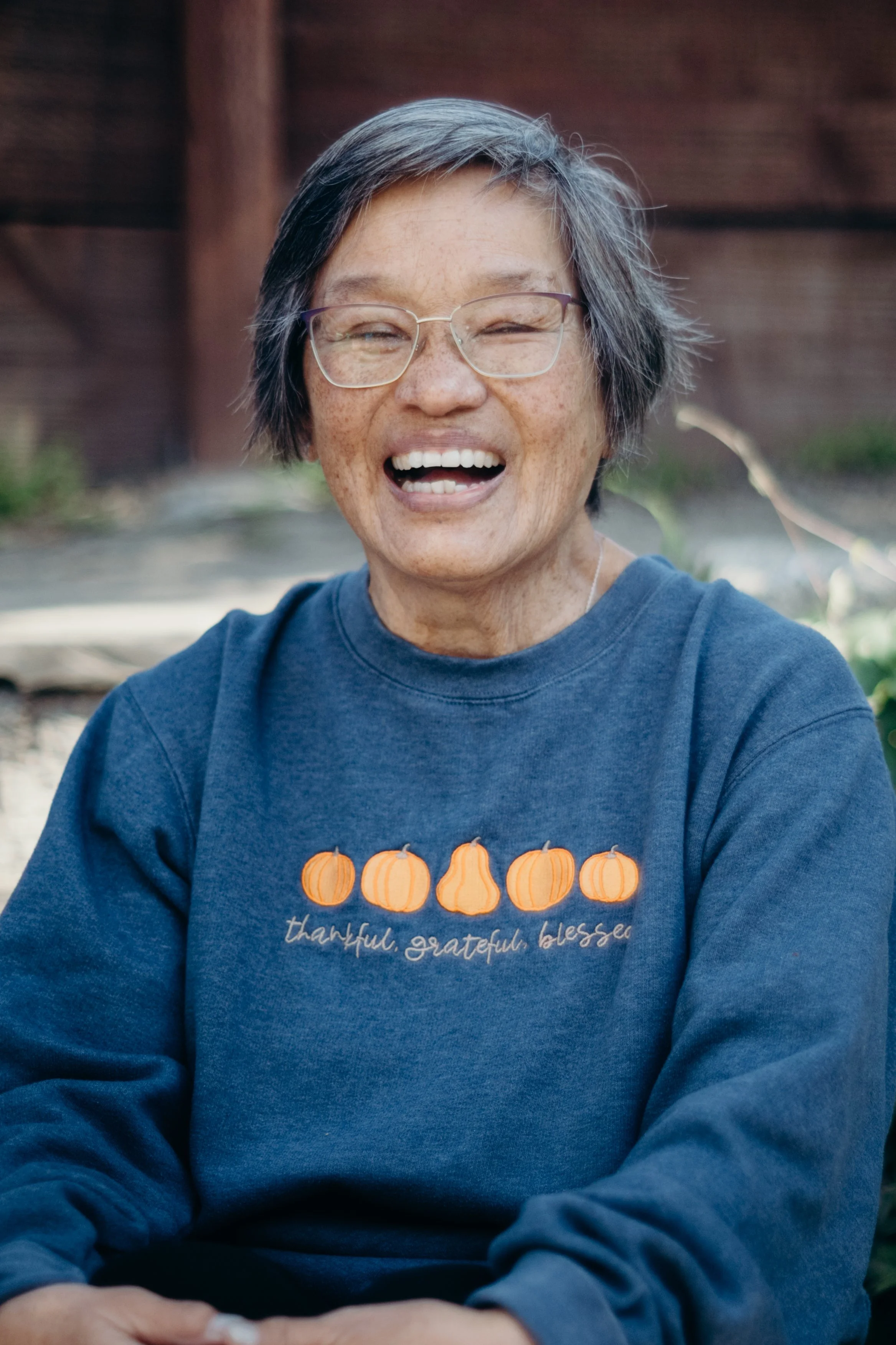 A woman with short gray hair and glasses smiling outdoor, wearing a blue sweatshirt with pumpkins and the words 'thankful, grateful, blessed' embroidered on it.
