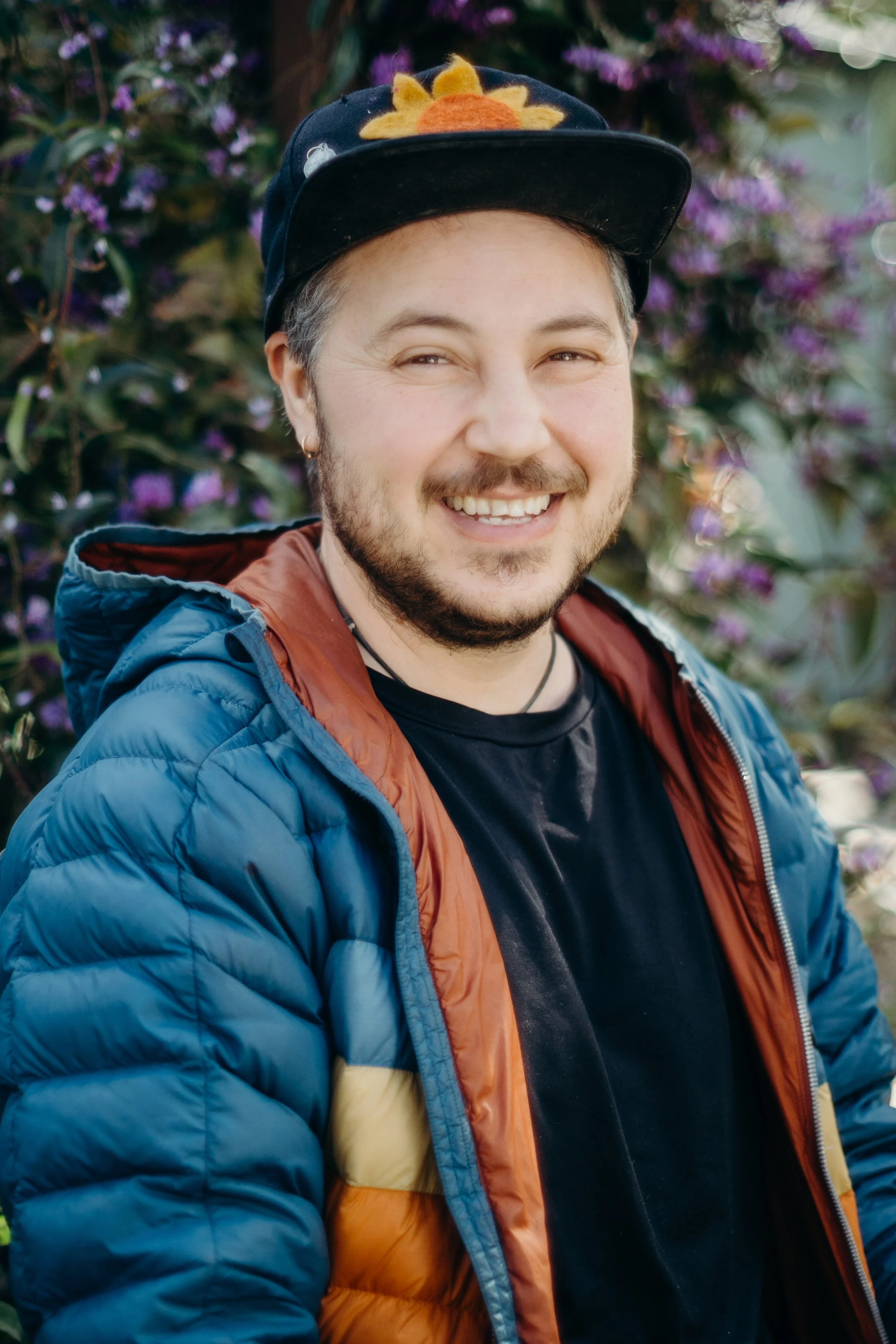 A smiling man with a beard and earrings wearing a baseball cap with a felt sunflower, a patchwork blue and orange puffer jacket, and a black t-shirt, standing outdoors with purple flowering bushes in the background.