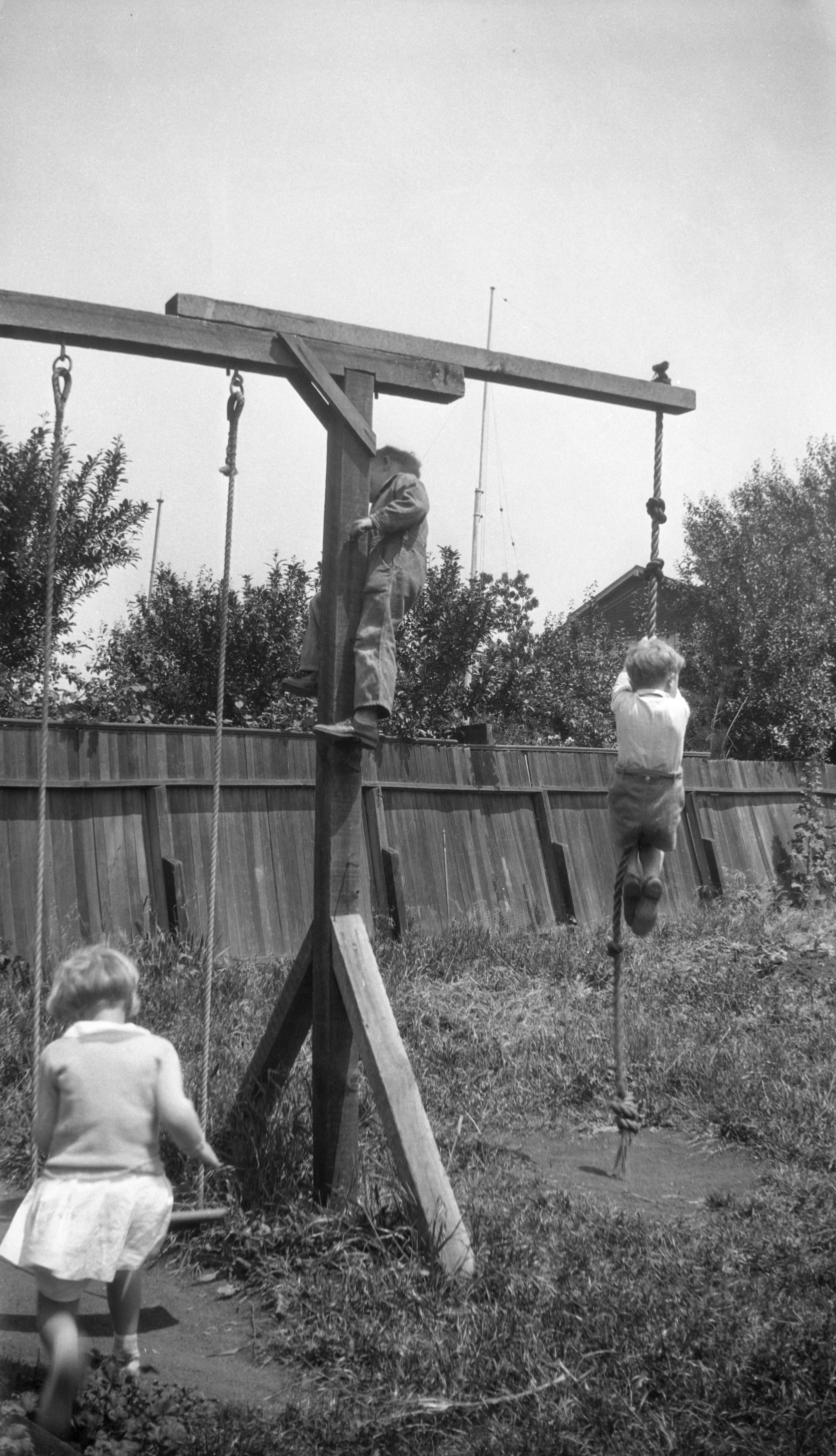 Children playing on a wooden outdoor playground set with swings, one boy climbing a vertical log and a girl on a rope swing, in a backyard with a wooden fence and trees.