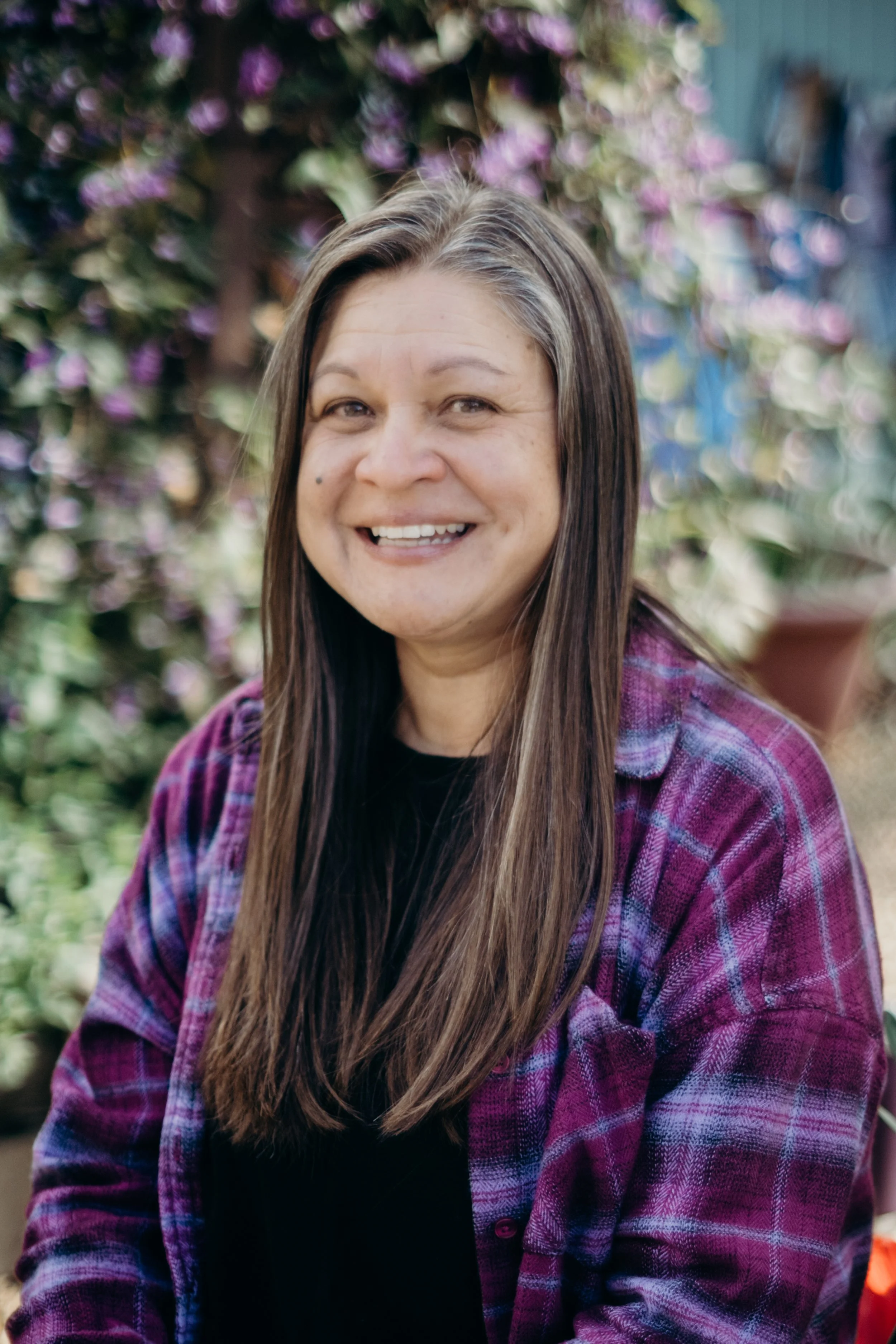 A woman with long brown hair, smiling, wearing a purple and black plaid shirt and a black top, outside with plants and flowers in the background.