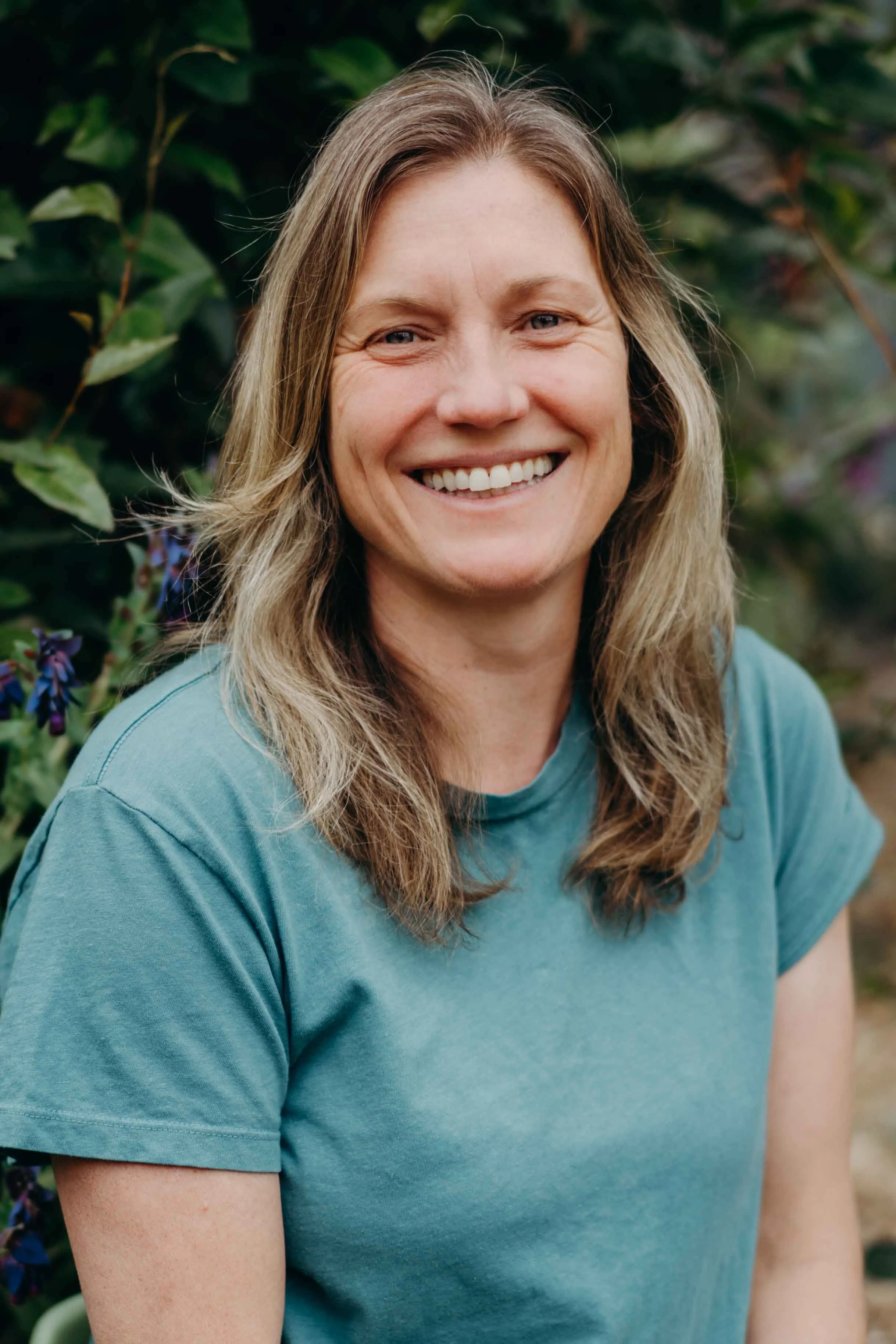 A woman with light brown, wavy hair smiling outdoors in front of green foliage and purple flowers, wearing a blue t-shirt.