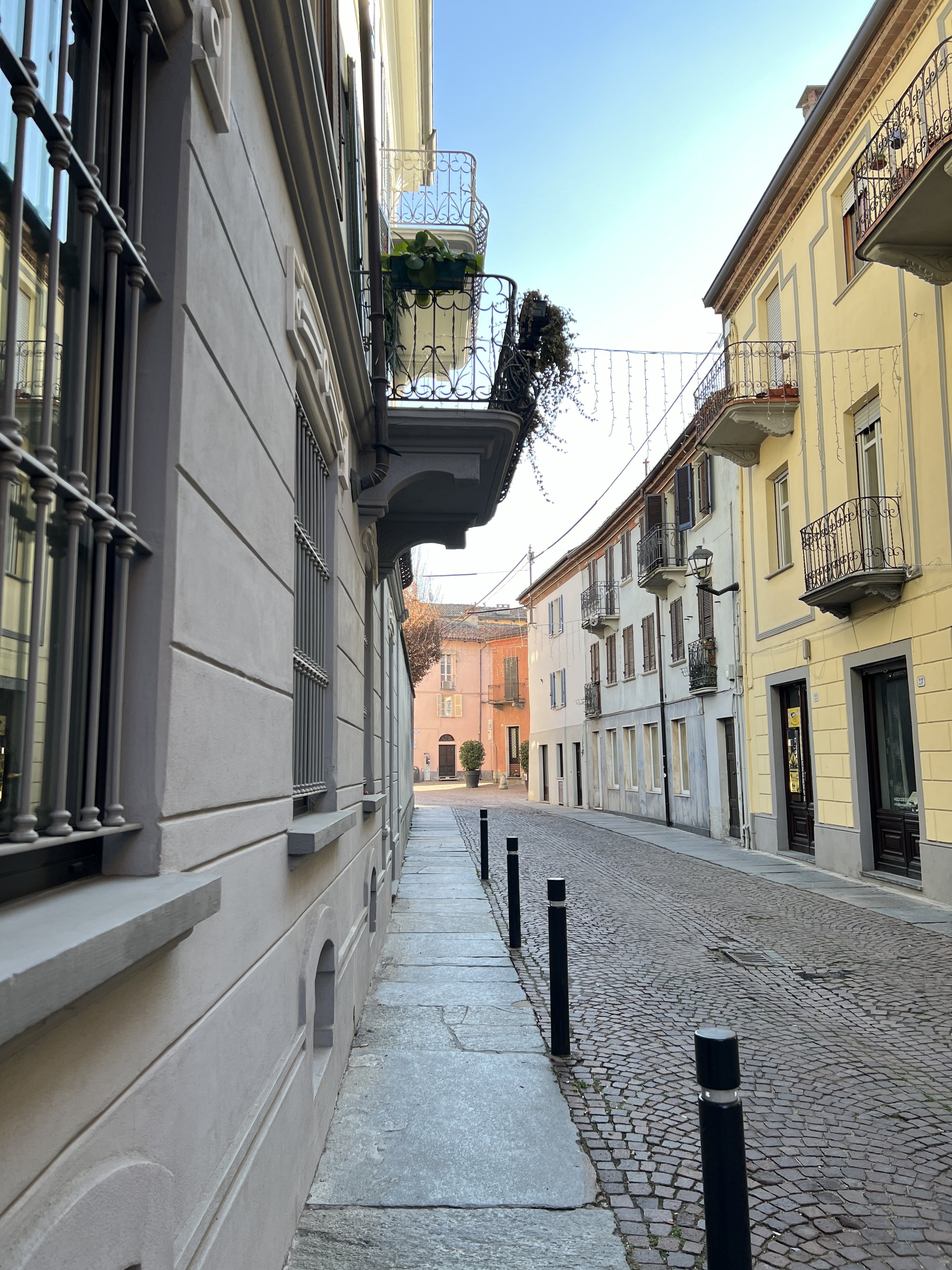 A narrow cobblestone street in a European city, lined with pastel-colored buildings with balconies and small windows, during daytime with clear blue sky.