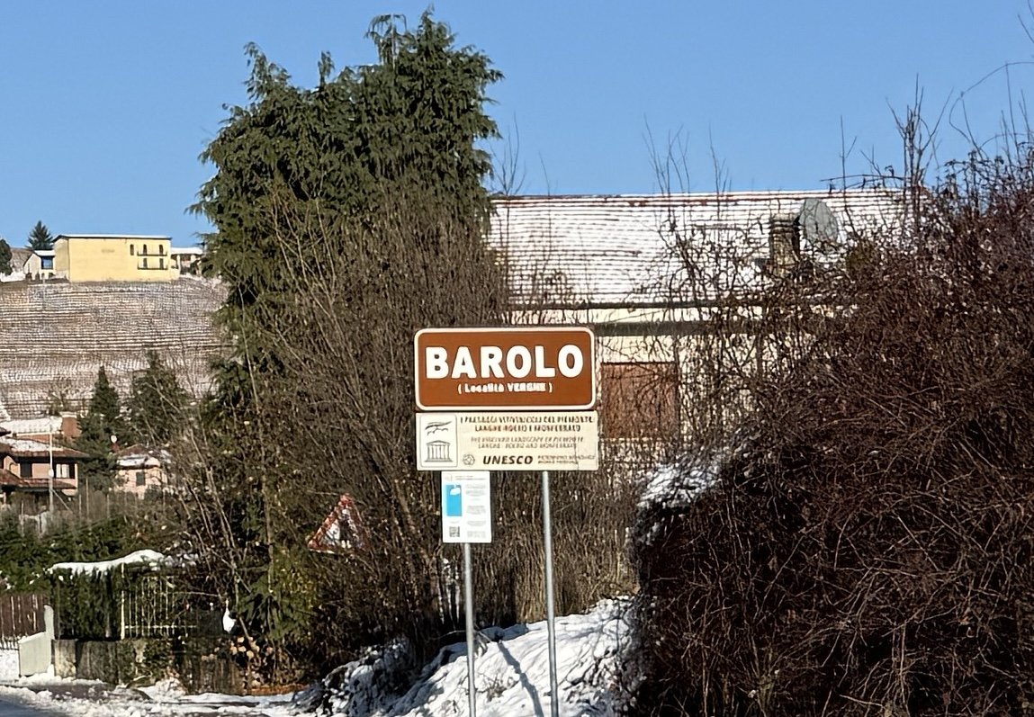 Sign indicating the entrance to Barolo, Italy, with snow on the ground and bushes around.