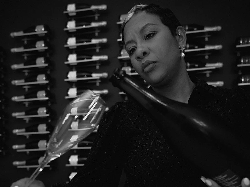 A woman with short hair and pearl earrings is looking at a wine bottle while holding a wine glass, standing in front of a wall decorated with arranged wine bottles.