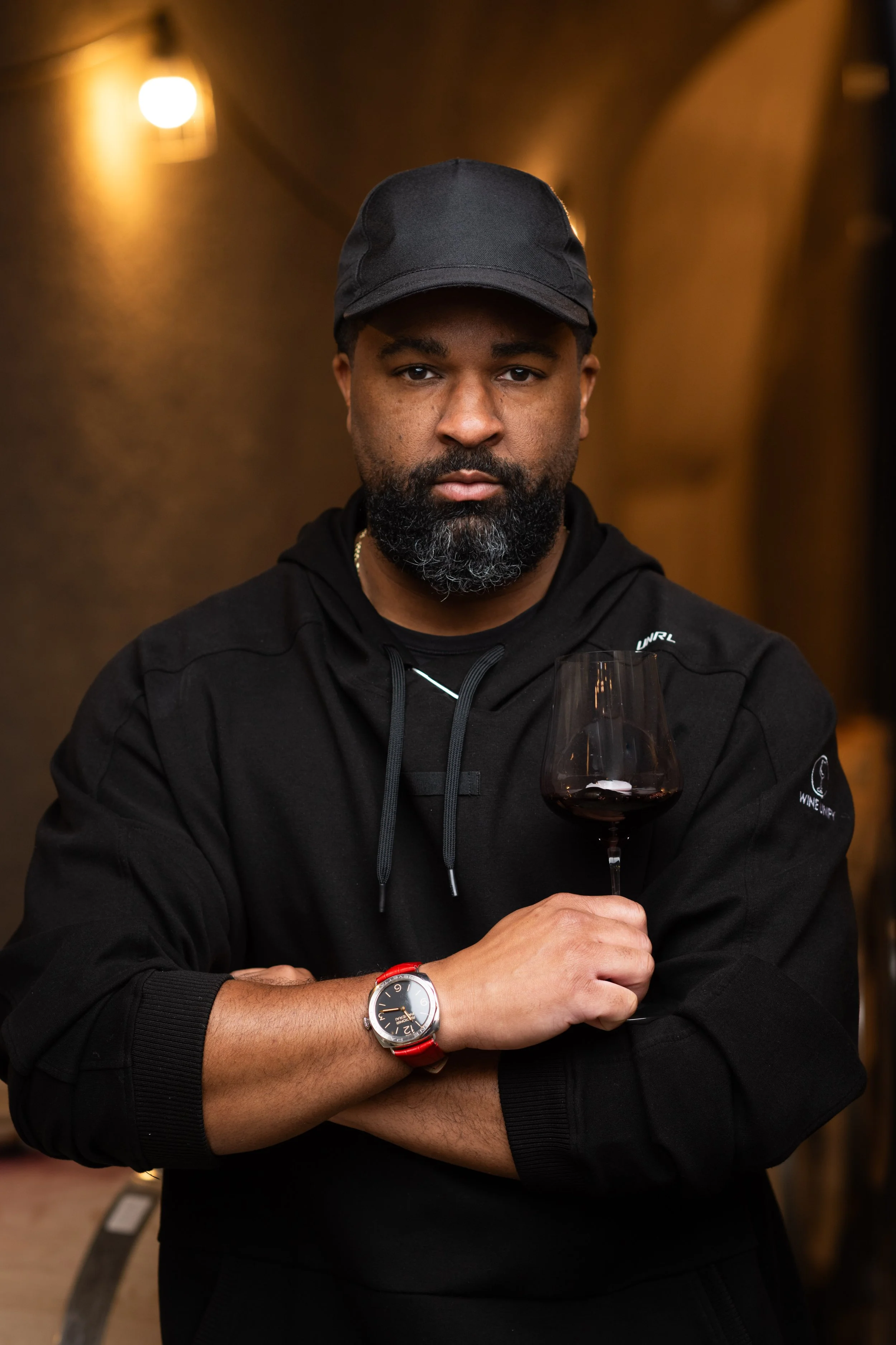 DLynn Proctor, wearing a black cap and black wine unify hoodie, holds a glass of cabernet red wine in his right hand, standing in a dimly lit cellar.