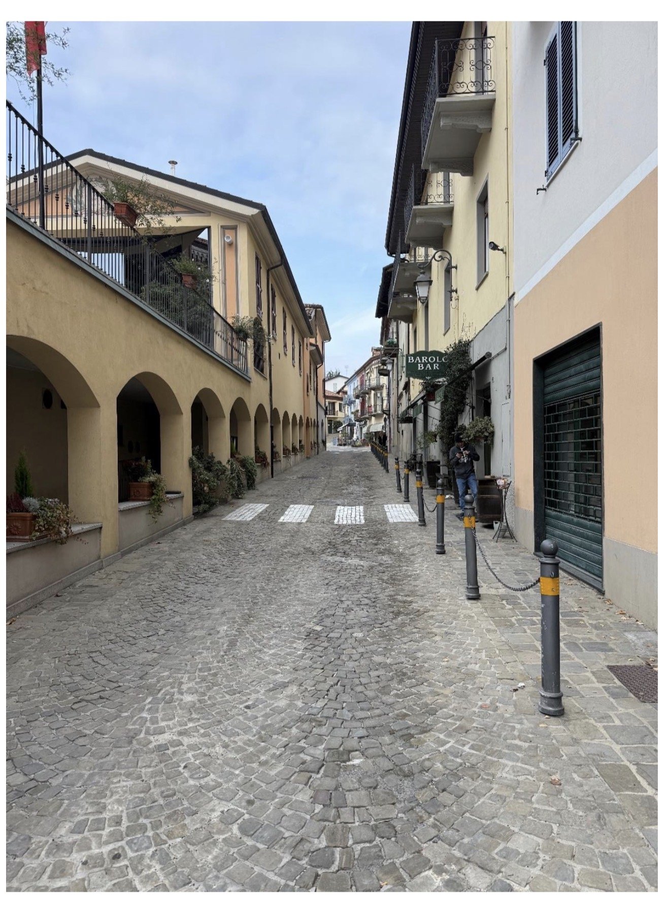 A narrow cobblestone street in a European town, lined with pastel-colored buildings, some with balconies and shutters. There are bollards along the sidewalk, and a sign for Barolo Bar visible on the right side.