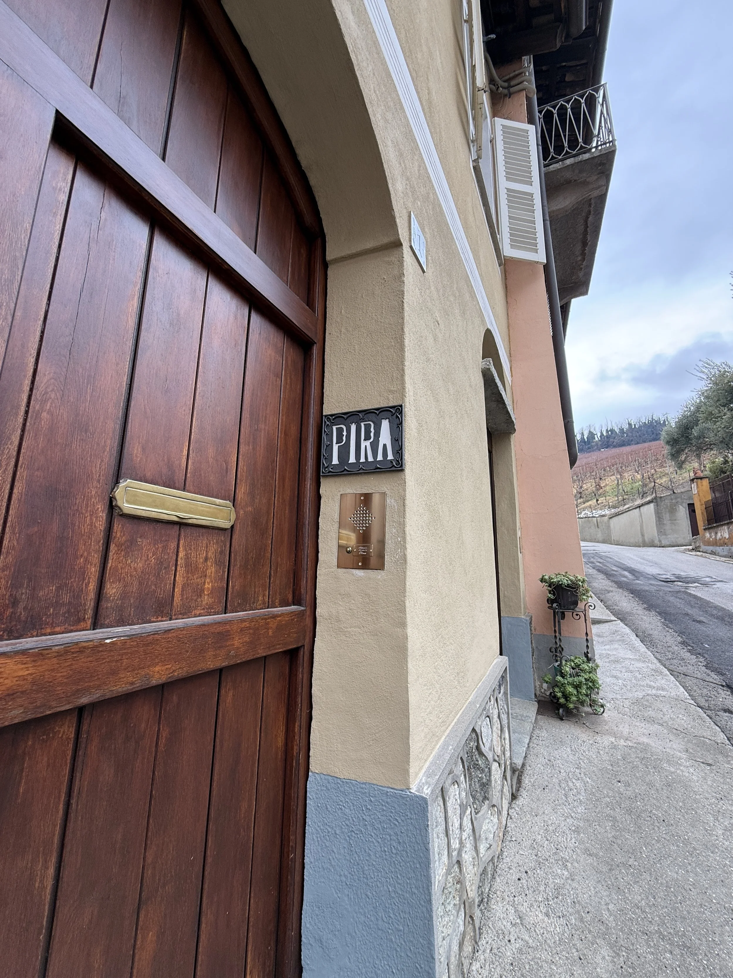 A close-up of a wooden garage door on a beige house, with a decorative sign that says 'PIRA' and a doorbell below it. The sidewalk and street are visible to the right, with a potted plant near the corner of the house.