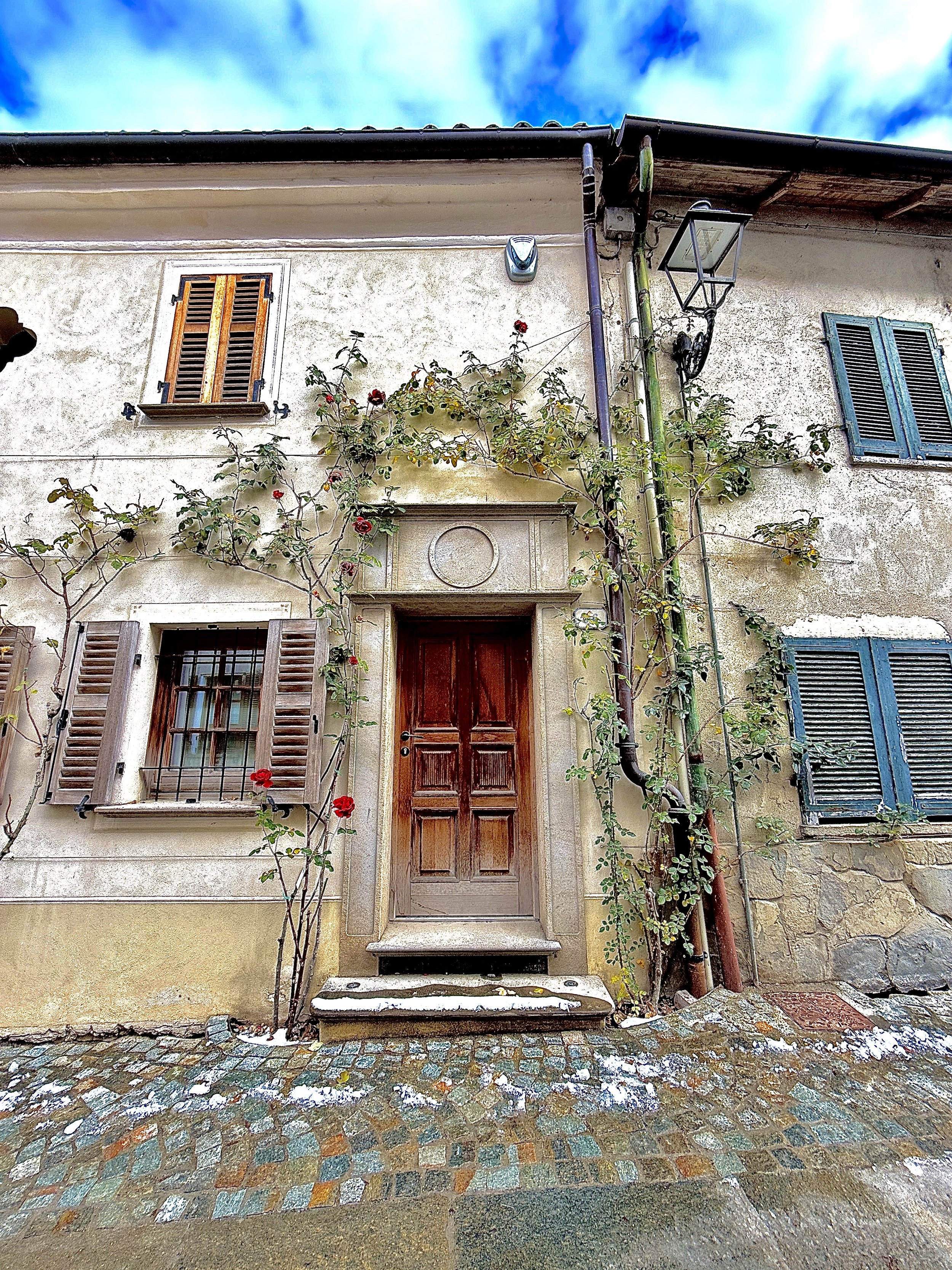 A rustic exterior of a house with a wooden door, two windows with shutters, some flowering climbing plants, and cobblestone ground in Bossolasco.