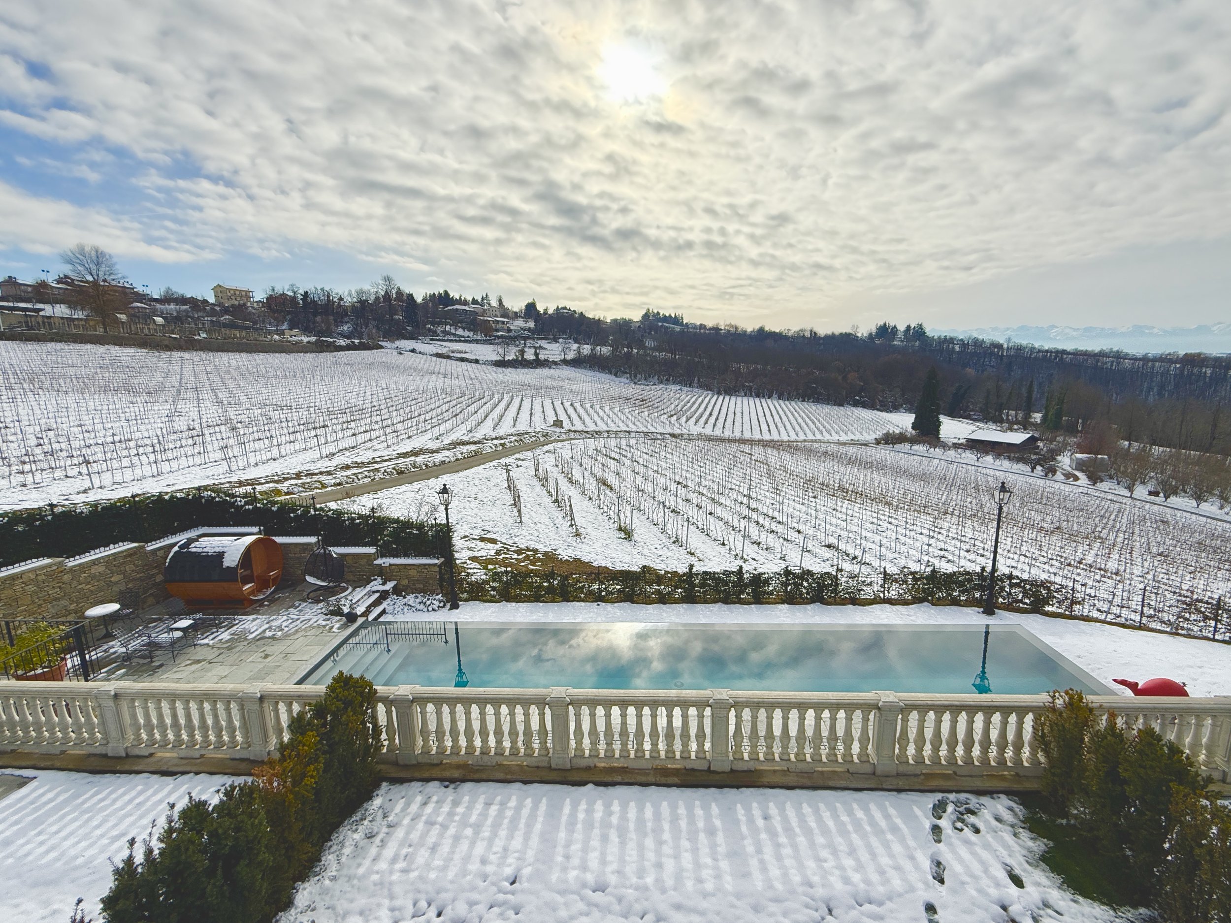 Snow-covered vineyard and hills with cloudy sky above, viewed from a backyard pool area with a white railing and snow-covered ground.