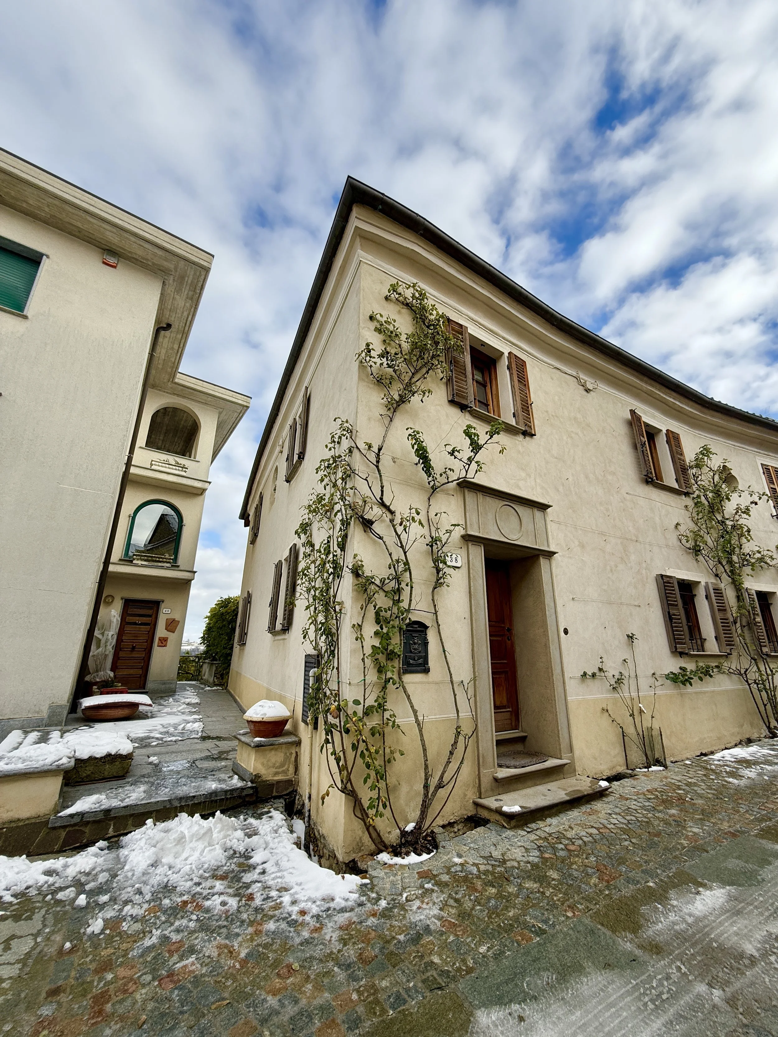 A cream-colored house with wooden shutters, climbing plants, and a snow-covered cobblestone street, with a cloudy blue sky.