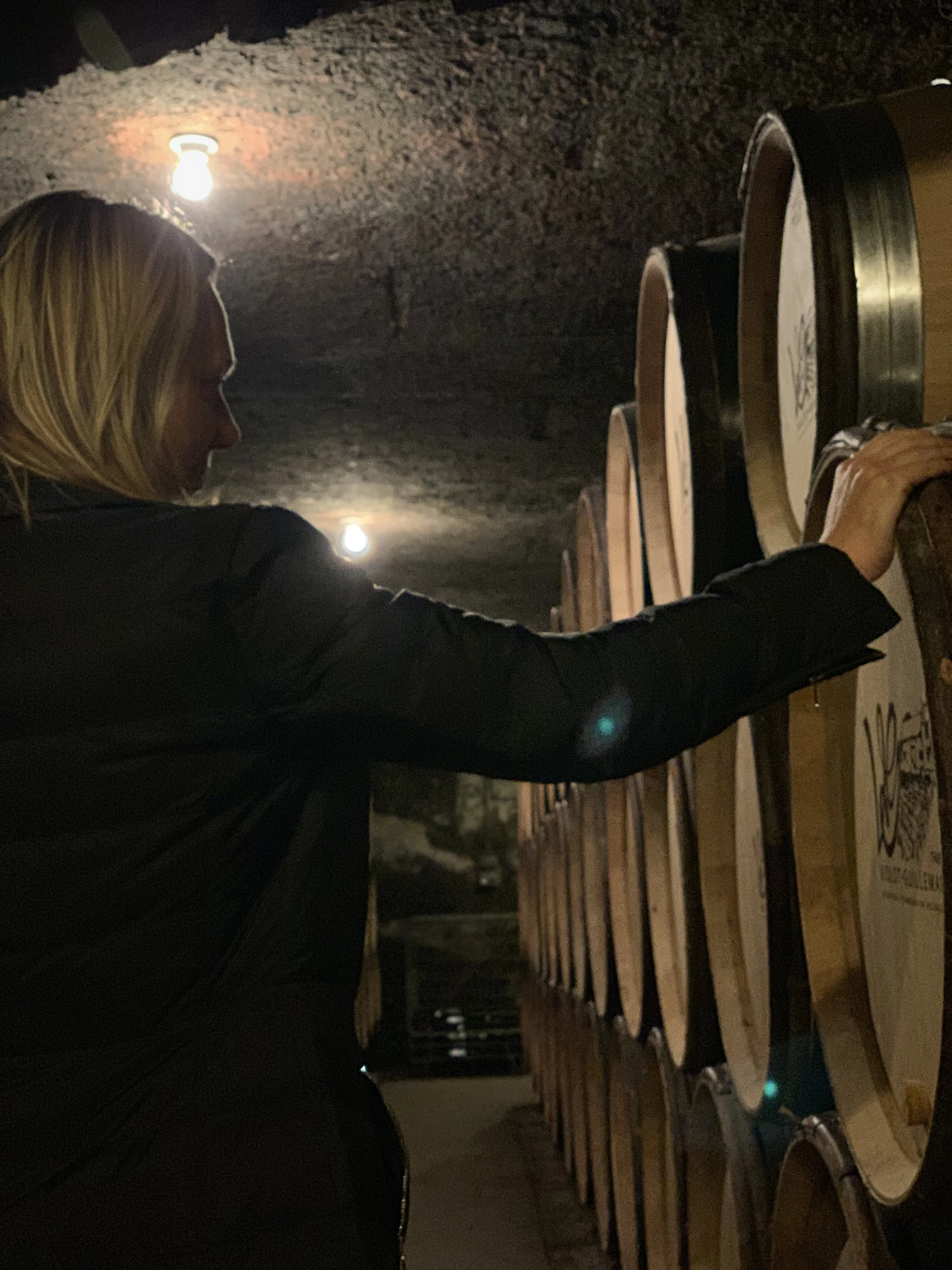A woman with blonde hair in a black jacket reaching out to touch wine barrels in a cellar or winery.