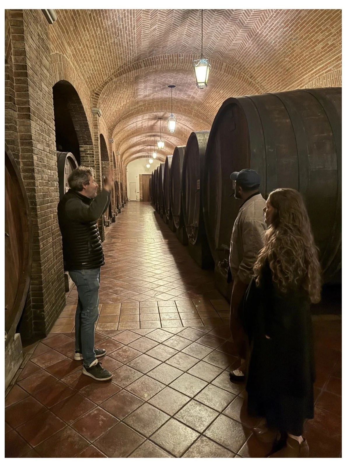 A wine cellar with large wooden barrels along the wall, three people engaged in conversation, with brick arched ceiling and warm lighting at Giacomo Conterno.