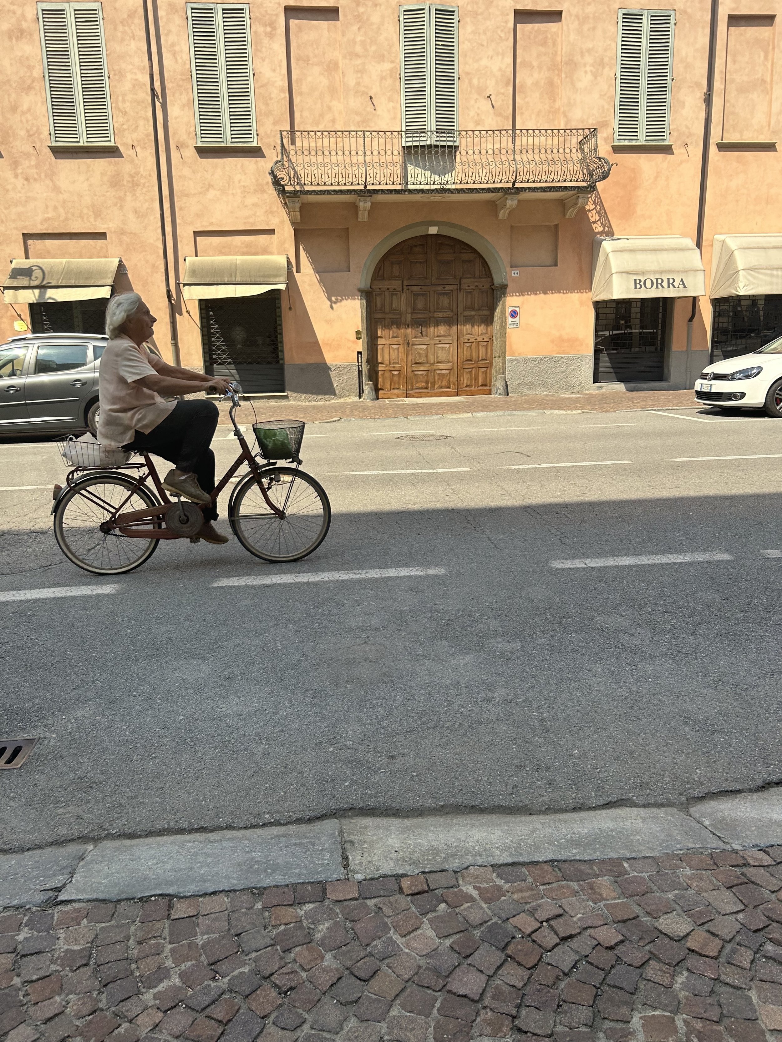 An elderly woman riding a bicycle on a city street with an orange building in the background and parked cars, in Cherasco.