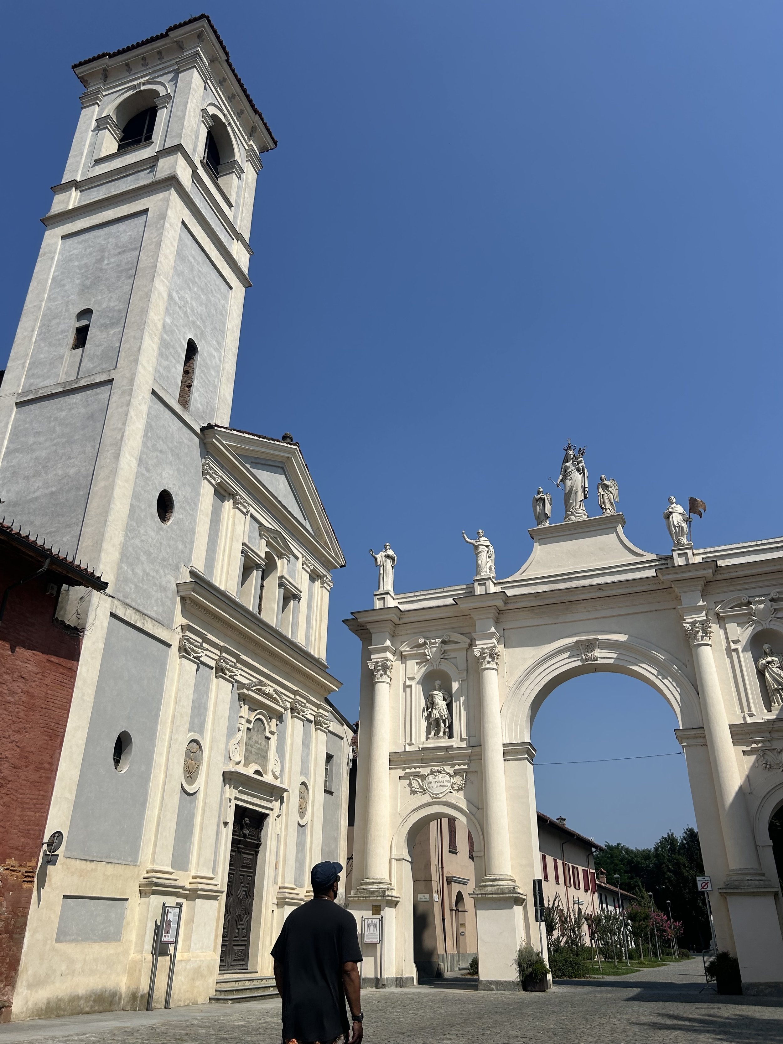 A man standing in front of a historic church with a tall bell tower, ornate statues, and a grand archway entrance under a clear blue sky.
